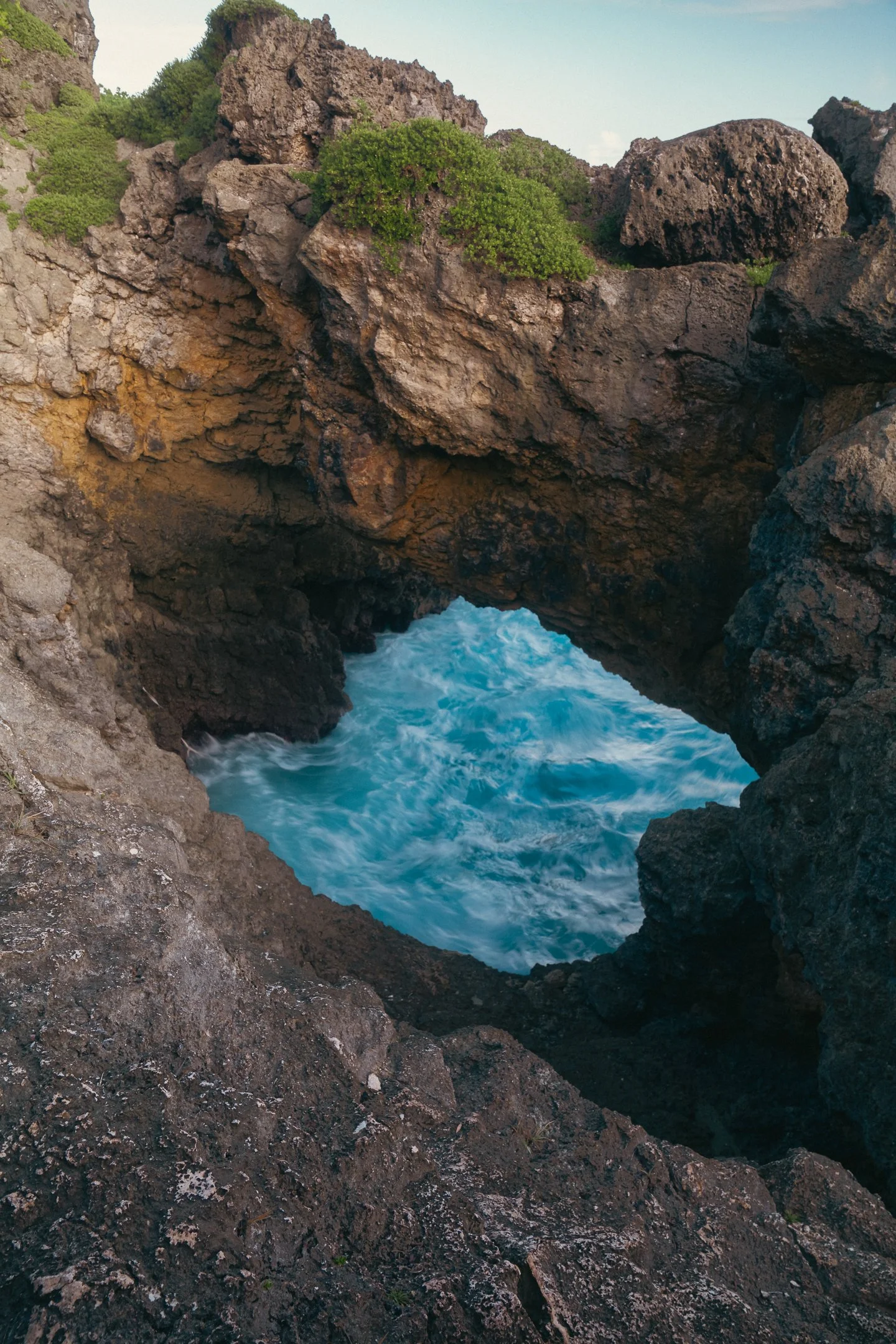 Ocean waves crashing through a natural rock formation with an arch, surrounded by rugged volcanic rocks with some green foliage on top.