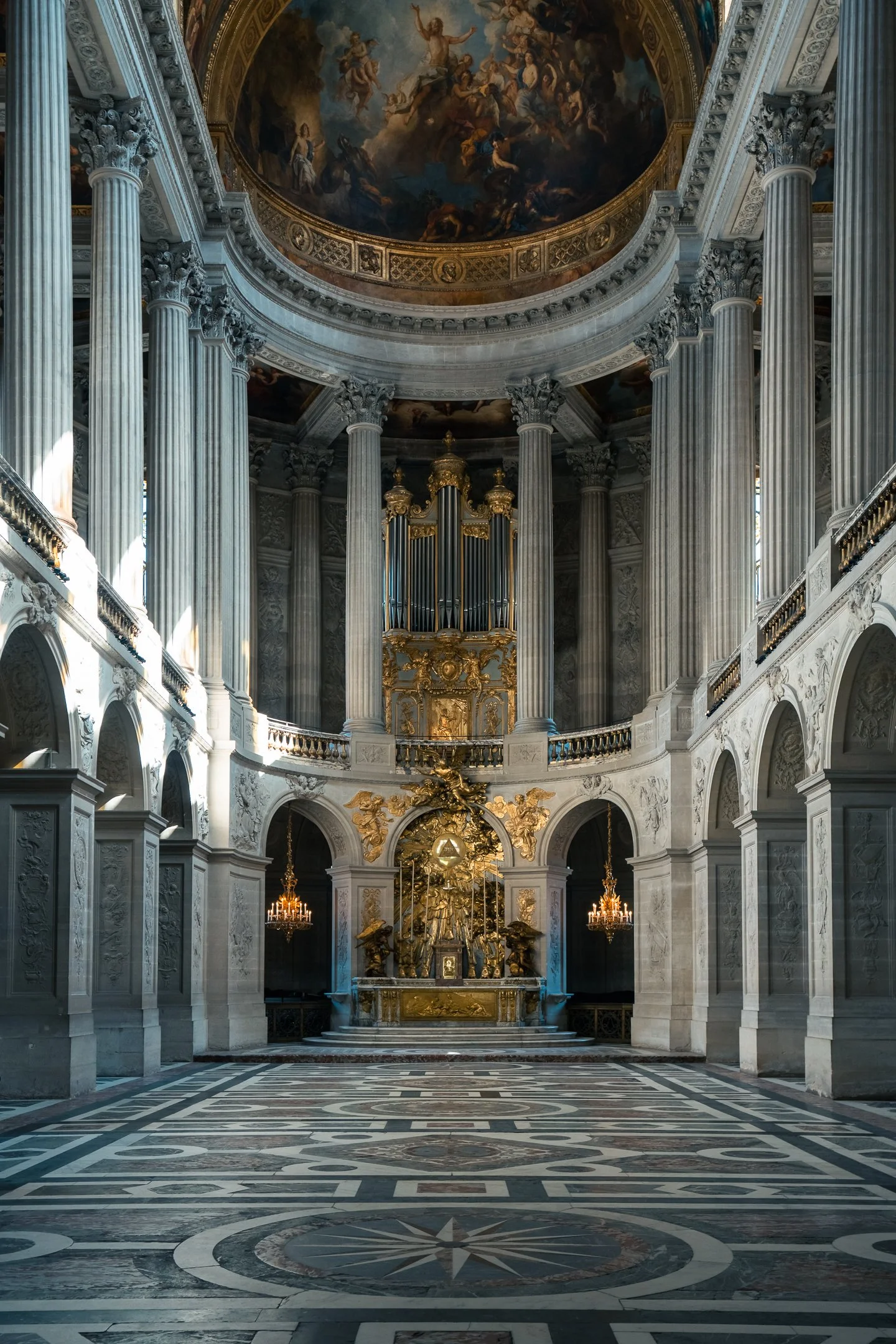 Interior of a grand, ornate church or cathedral featuring tall, fluted columns, intricate gold detailing, chandeliers, and a domed ceiling with a mural painting.