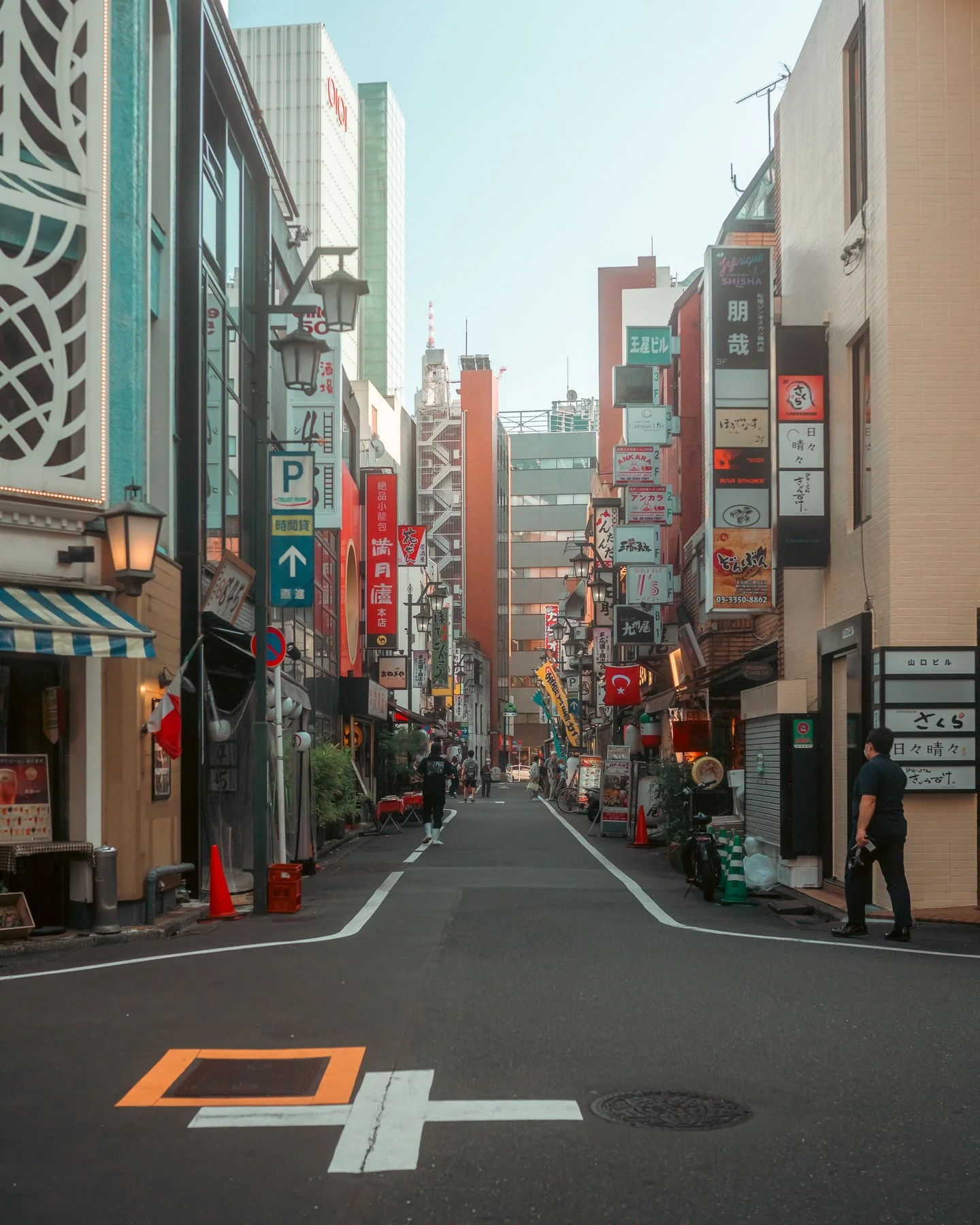 A narrow city street in Japan with tall buildings on both sides, colorful signs and banners in Japanese, a few pedestrians walking, and some outdoor seating areas.