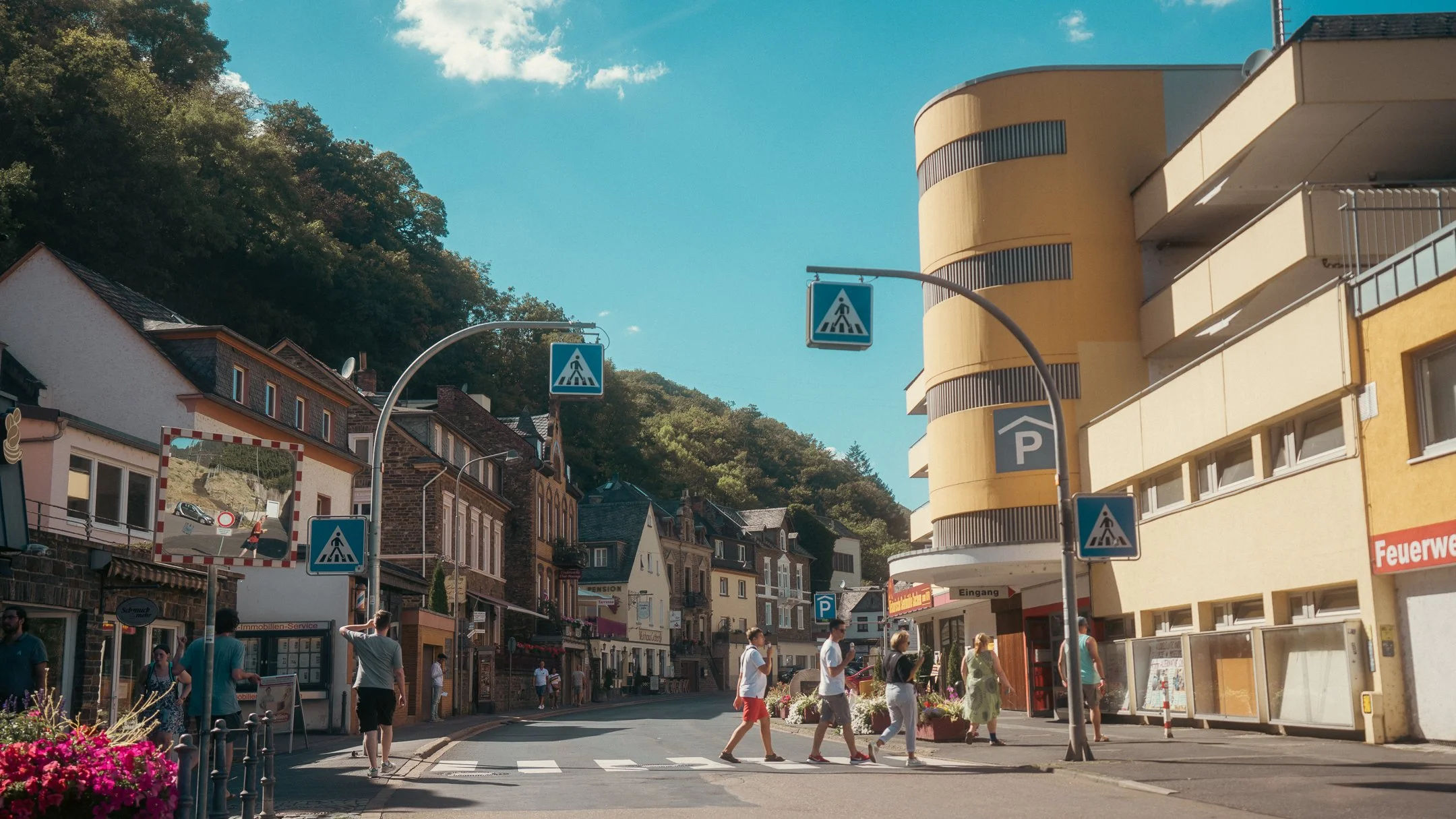 A crosswalk on a busy street in a small town with people walking and traffic signs, with buildings and a hillside in the background.