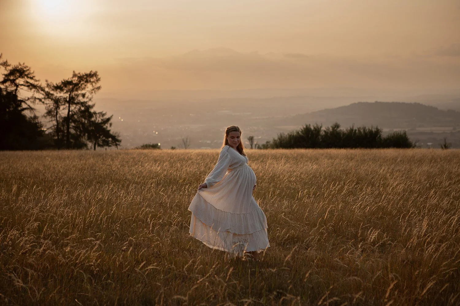 Pregnant woman in a linen ivory dress standing on a golden Cotswolds hill at sunset during an outdoor maternity photoshoot.