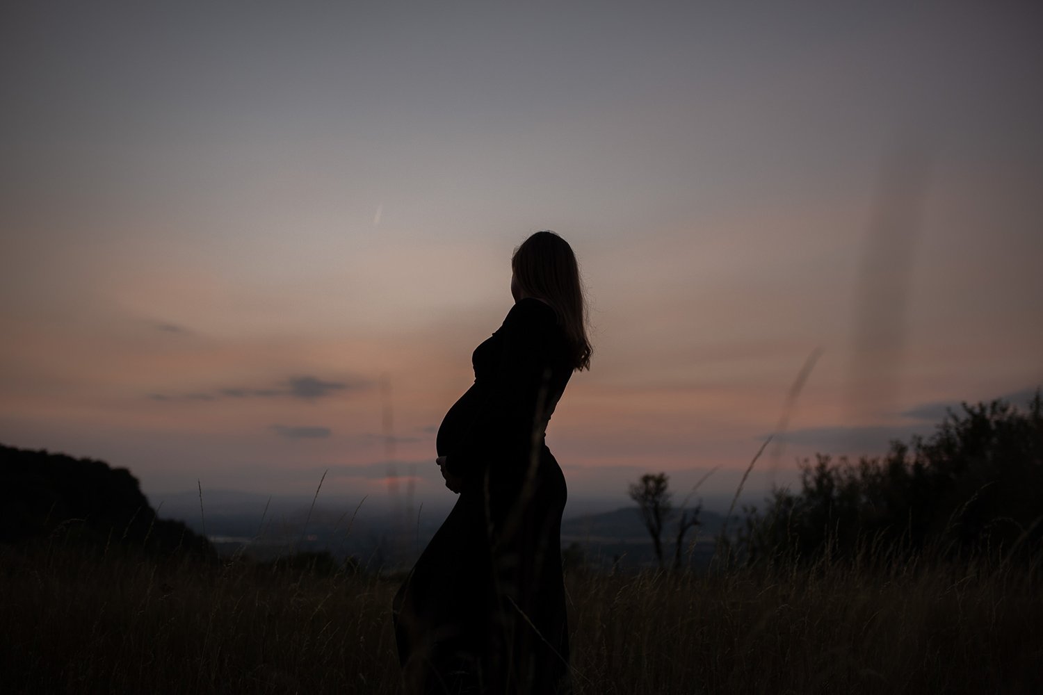 Pregnant woman in silhouette standing in a grassy Cotswolds hill at sunset, creating a artistic maternity portrait