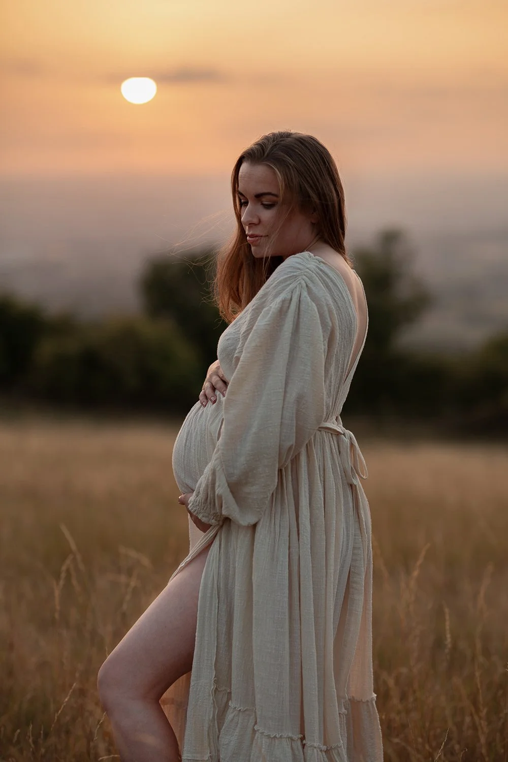 Pregnant woman in a linen cream dress standing in on a Cotswolds hill at sunset during an outdoor maternity photoshoot.