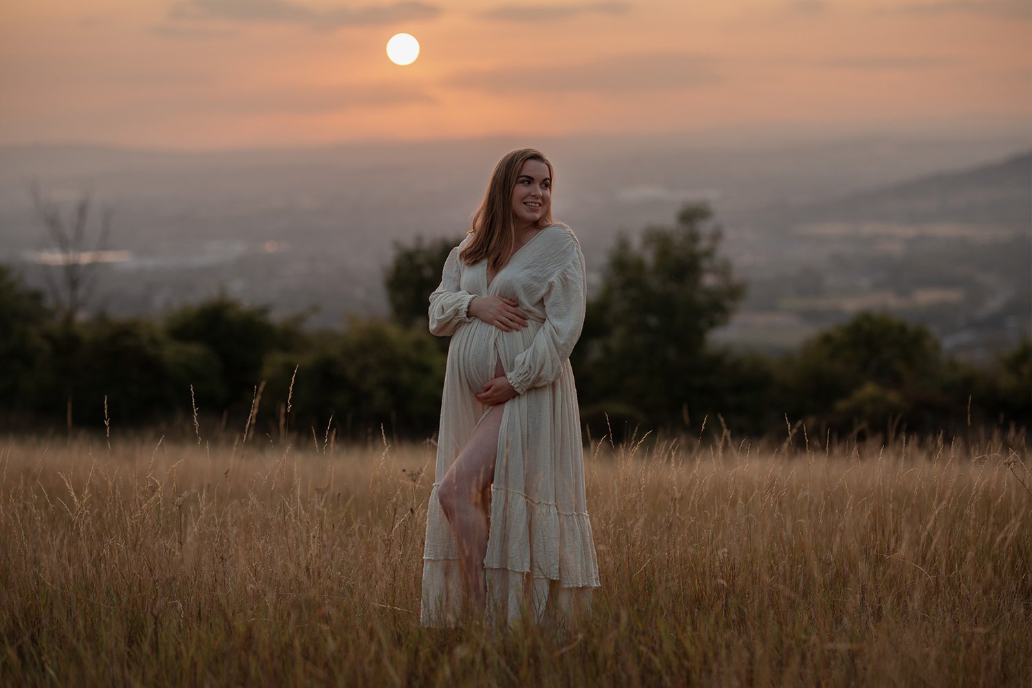 Pregnant woman in a linen ivory dress standing in a golden Cotswolds field at sunset during an outdoor maternity photoshoot.