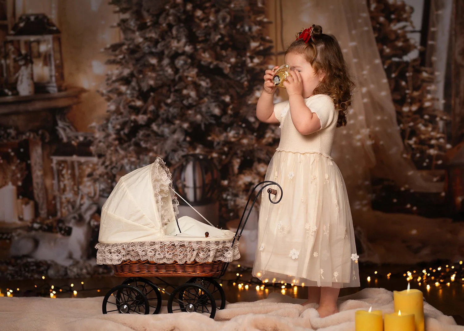 a young girl dressed in a white lace dress is playing with a toy camera pretending to take picture of a teddy in a toy pram during a Christmas mini session in Cheltenham