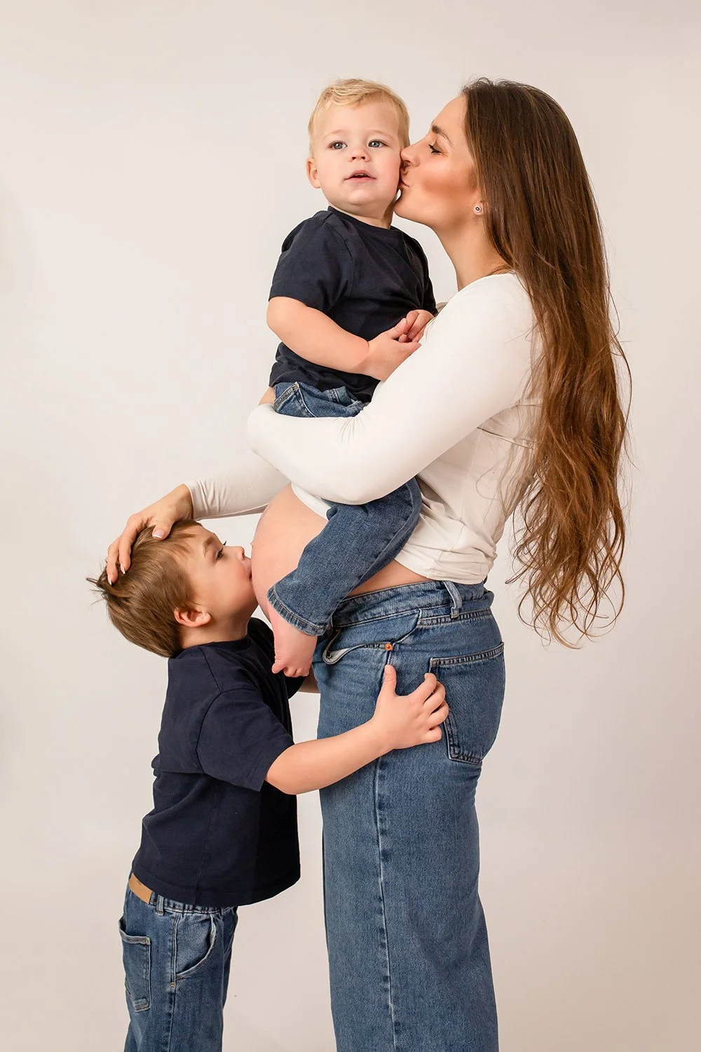 pregnant mum holding one toddler while the other one is kissing her bump during a maternity photoshoot in Cheltenham