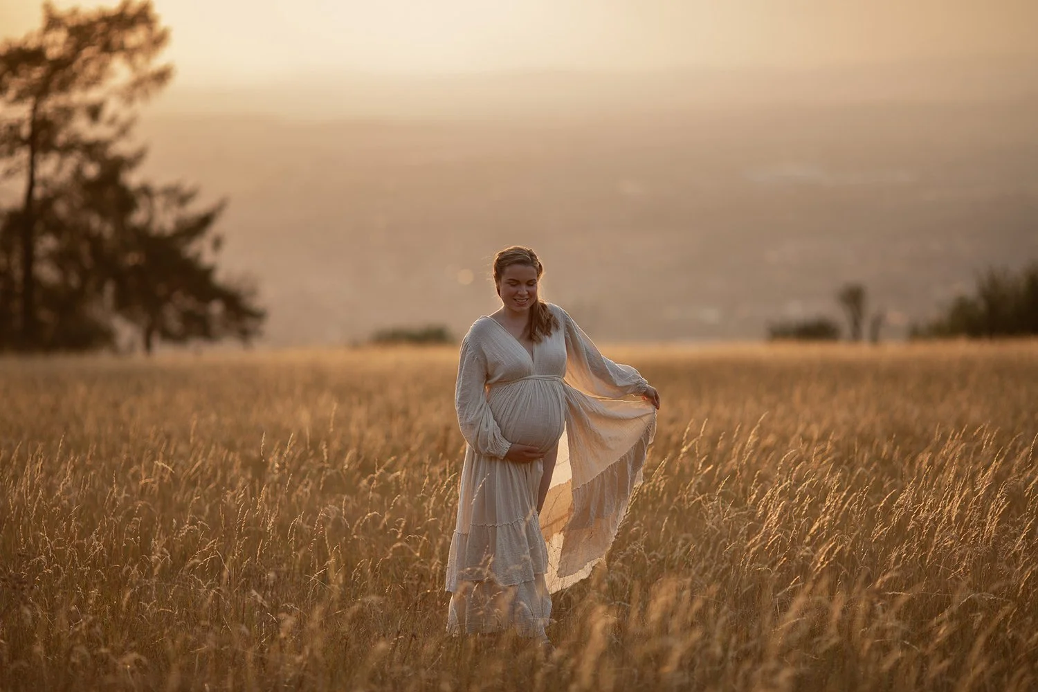 Pregnant woman in a linen dress walking in a golden Cheltenham field at golden hour during an outdoor maternity photoshoot.