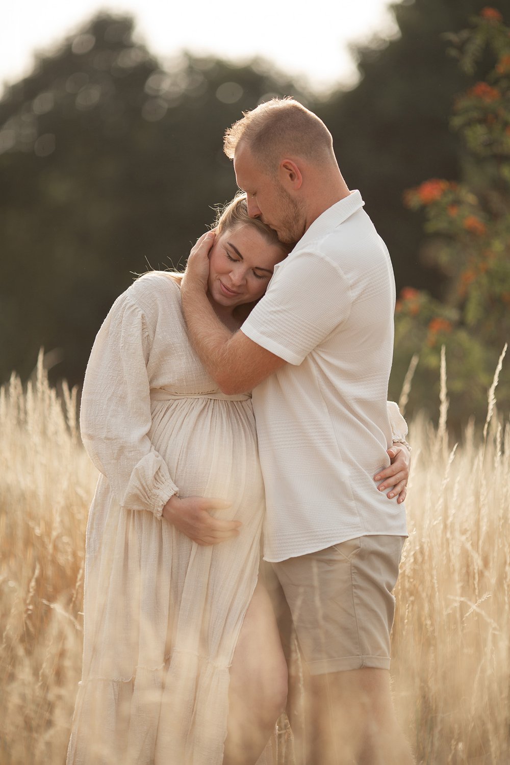 Expecting parents dressed in linen white and cream outfits cuddling during a summer outdoor maternity photoshoot in Cheltenham.