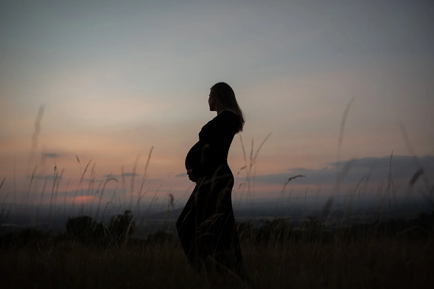 Pregnant woman in silhouette standing in a grassy Cotswolds field at sunset, creating a peaceful and atmospheric maternity portrait