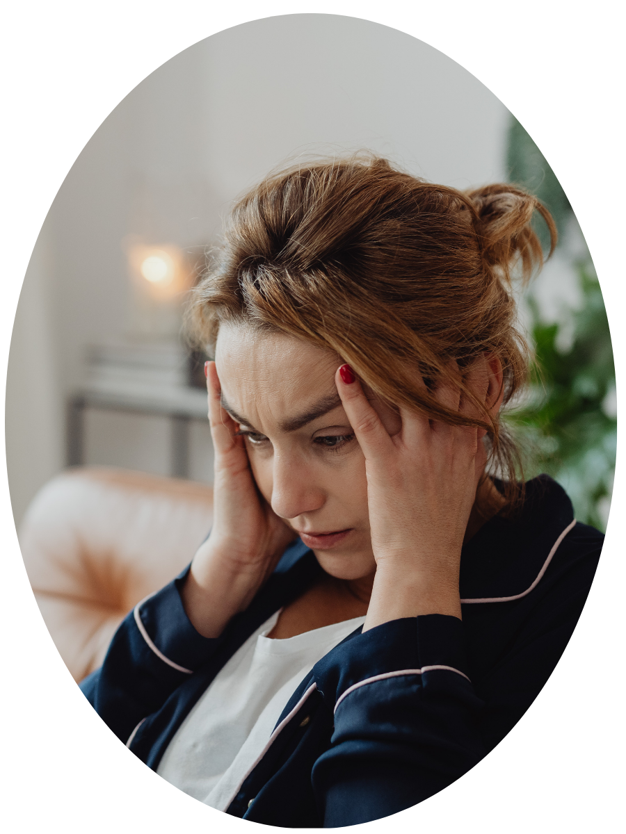 Woman with red hair holding her head, appearing stressed or worried, sitting on a beige couch in a living room.