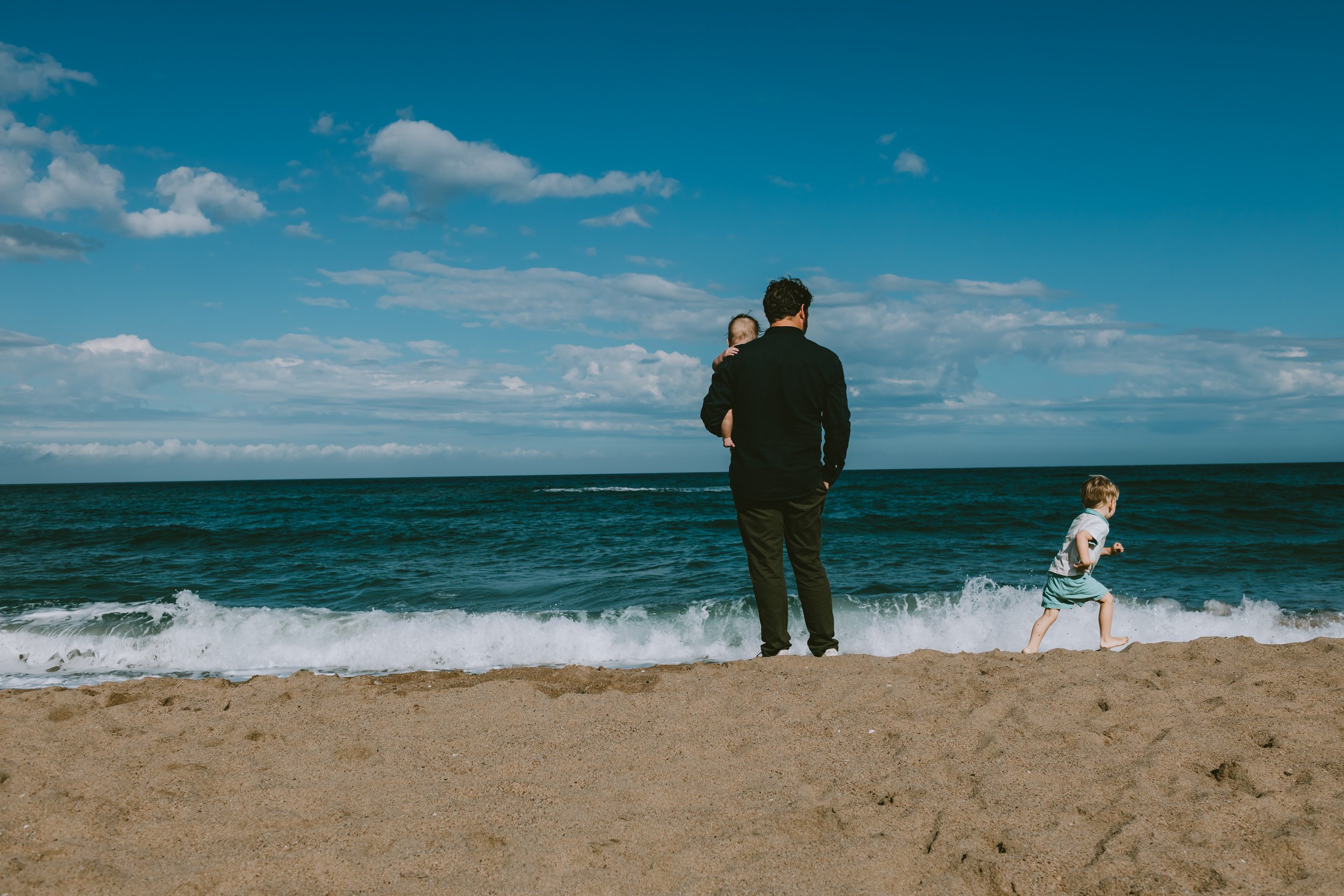 A man standing on a sandy beach holding a young child, and a small boy running towards the ocean waves under a blue sky with white clouds.