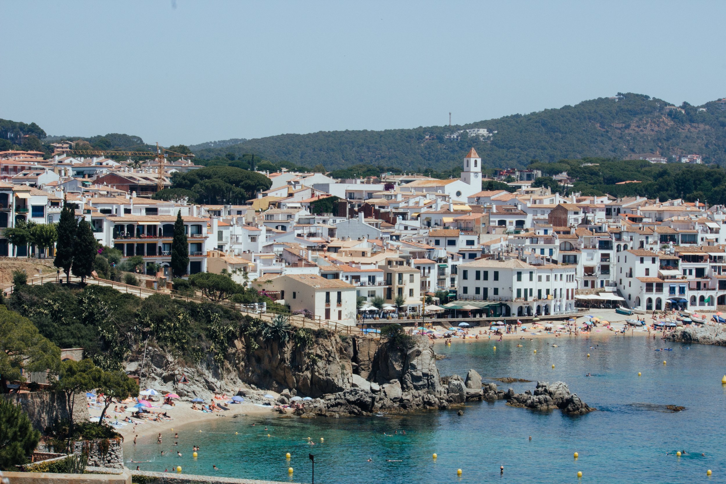 Coastal town with white buildings, a beach with umbrellas, and a calm turquoise sea.