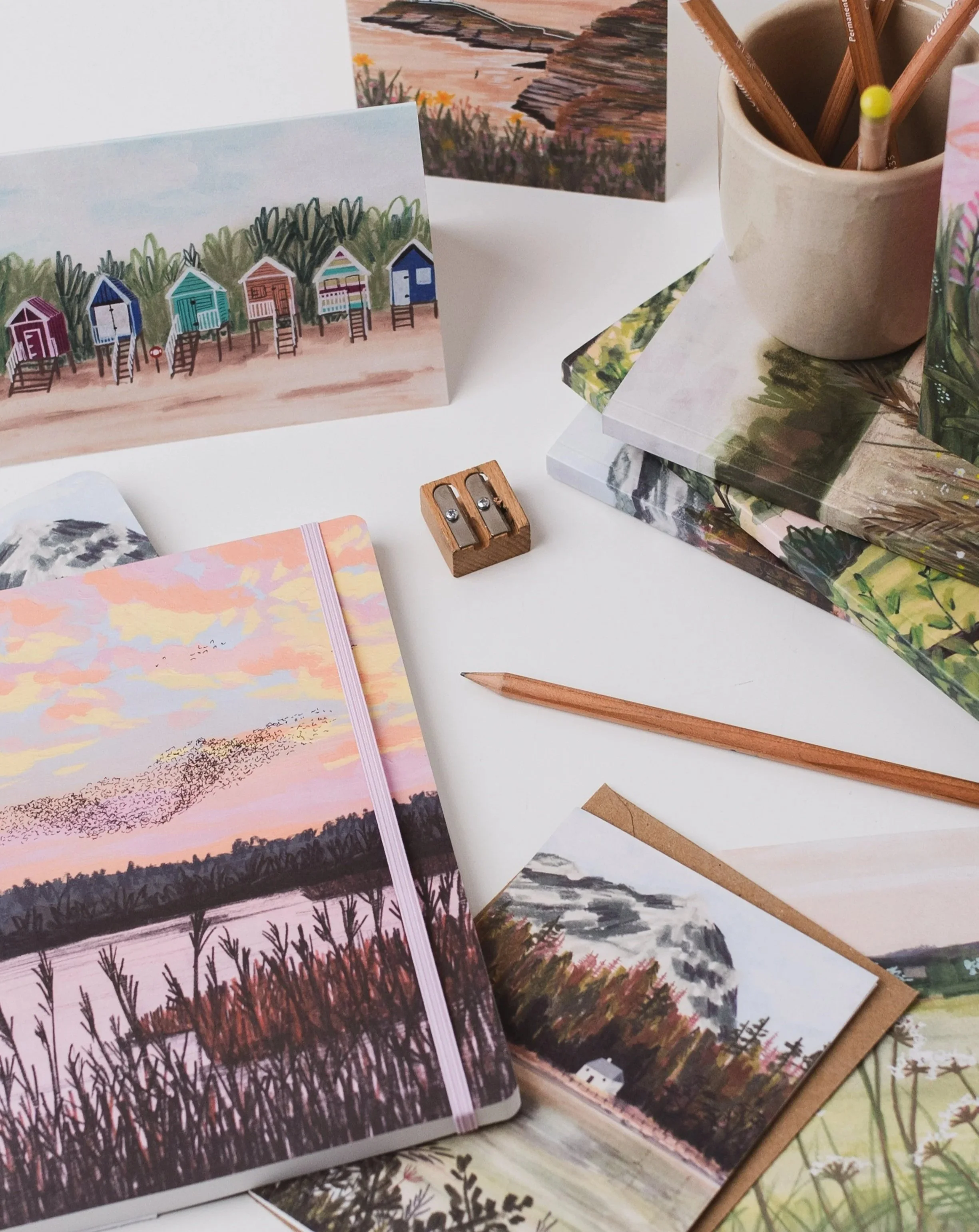 a flat lay of notebooks and greetings cards on a white desk, with some pencils and a sharpener