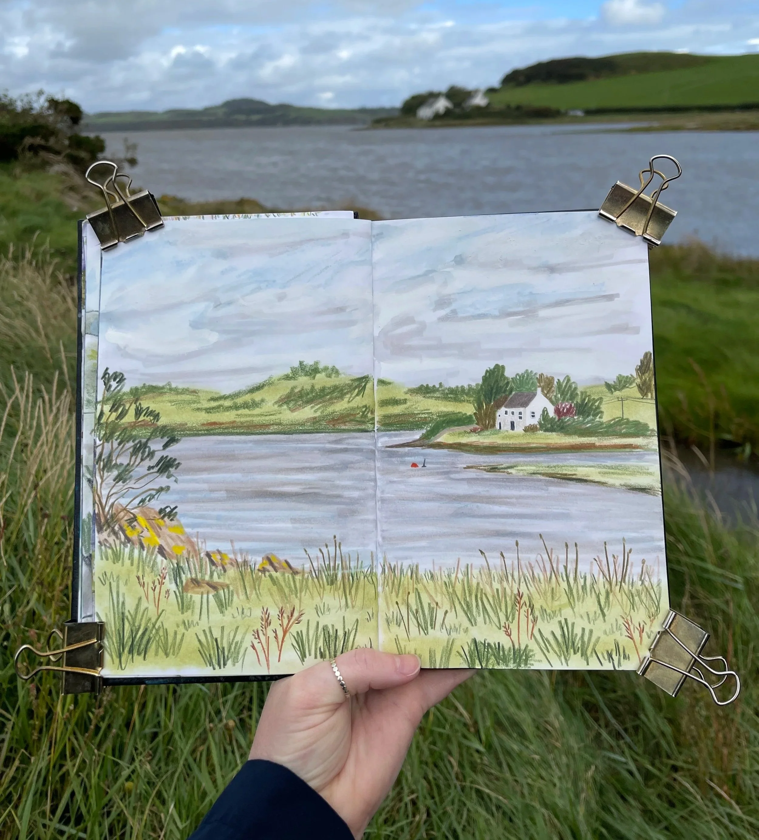 a sketchbook page with a landscape illustration being held in front of the landscape that inspired it