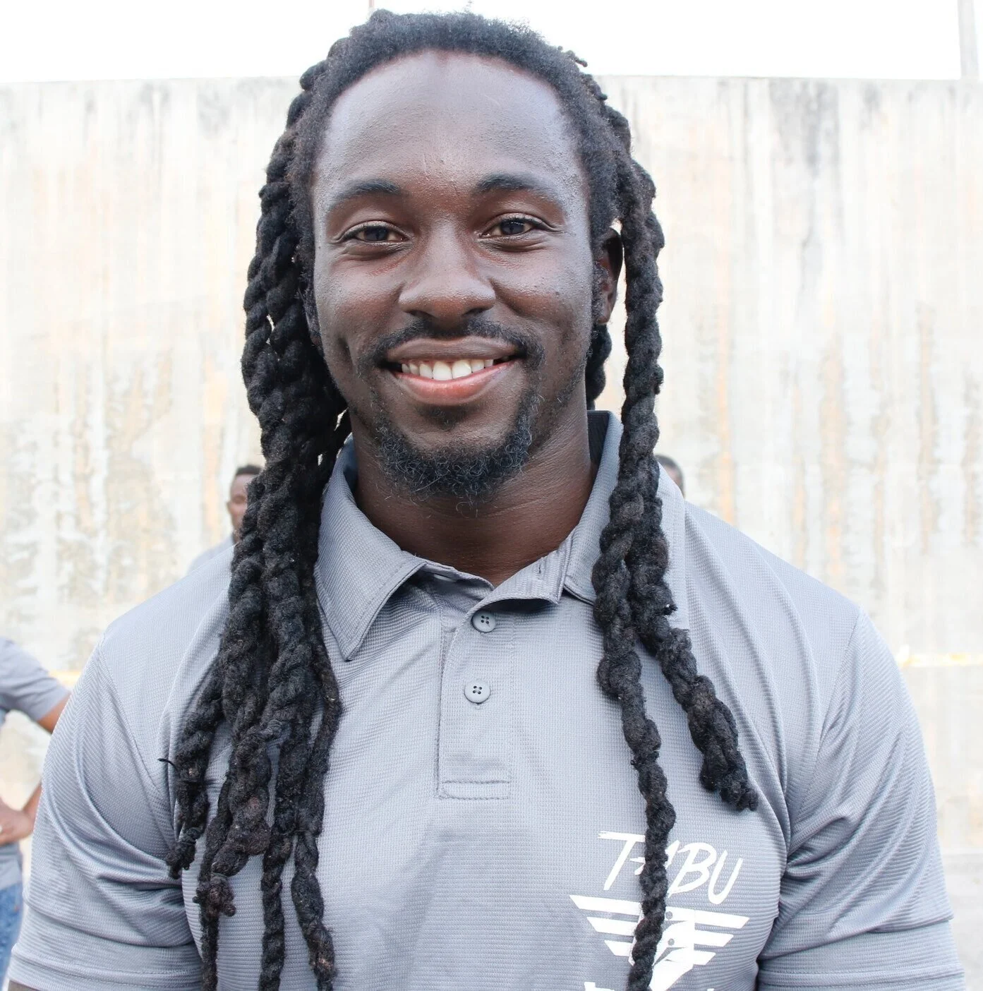 A smiling young man with long dreadlocks wearing a gray polo shirt with a logo, standing outdoors in front of a concrete wall.