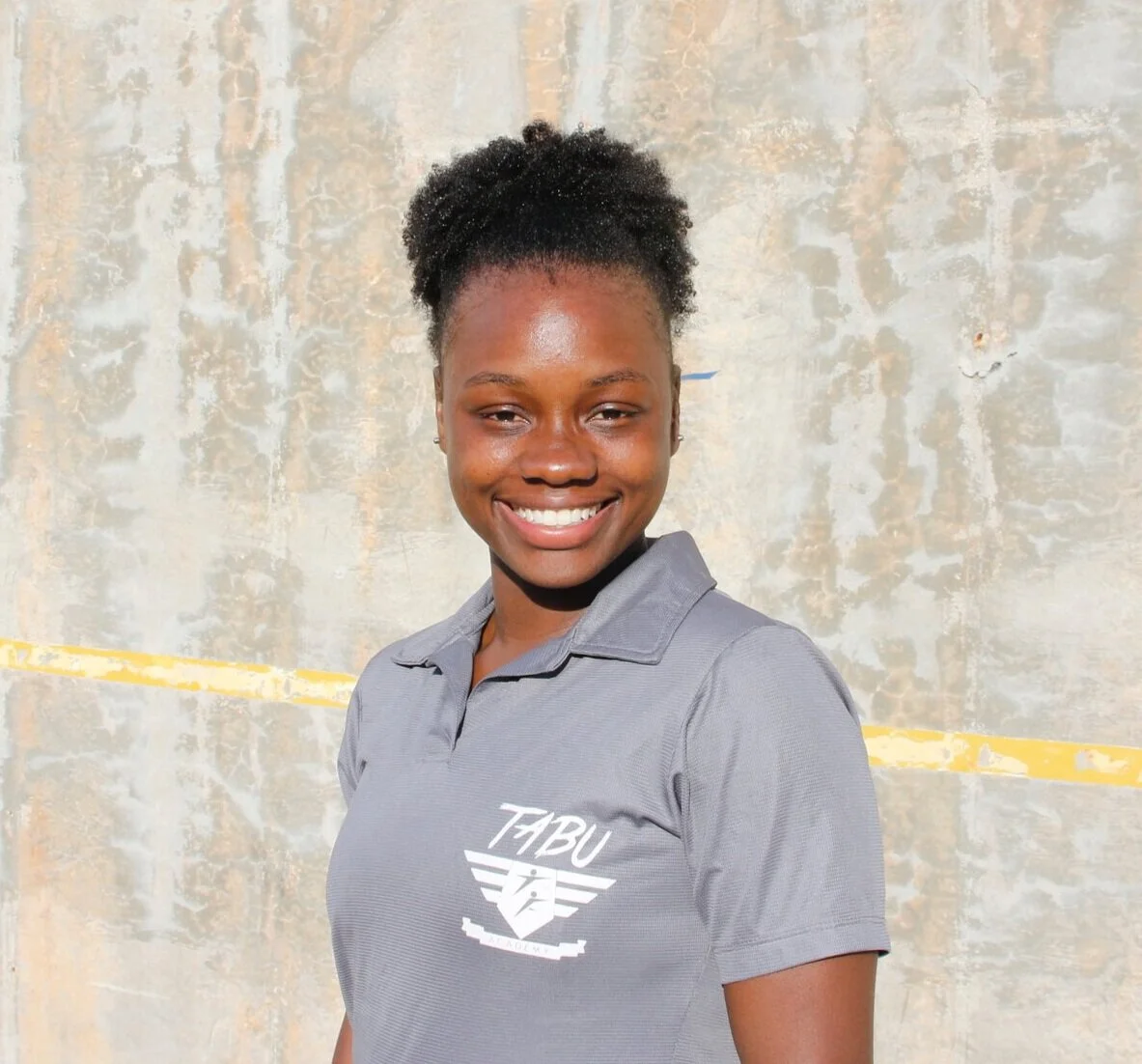 A young woman with dark skin and curly hair styled in a puff, smiling and wearing a gray polo shirt with a 'TABU' logo. She is standing in front of a weathered gray wall with yellow markings.