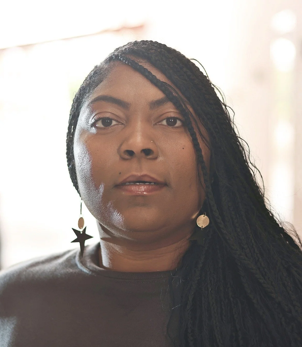 A woman with long braided hair wearing star-shaped earrings and a dark top, standing indoors with bright light behind her.