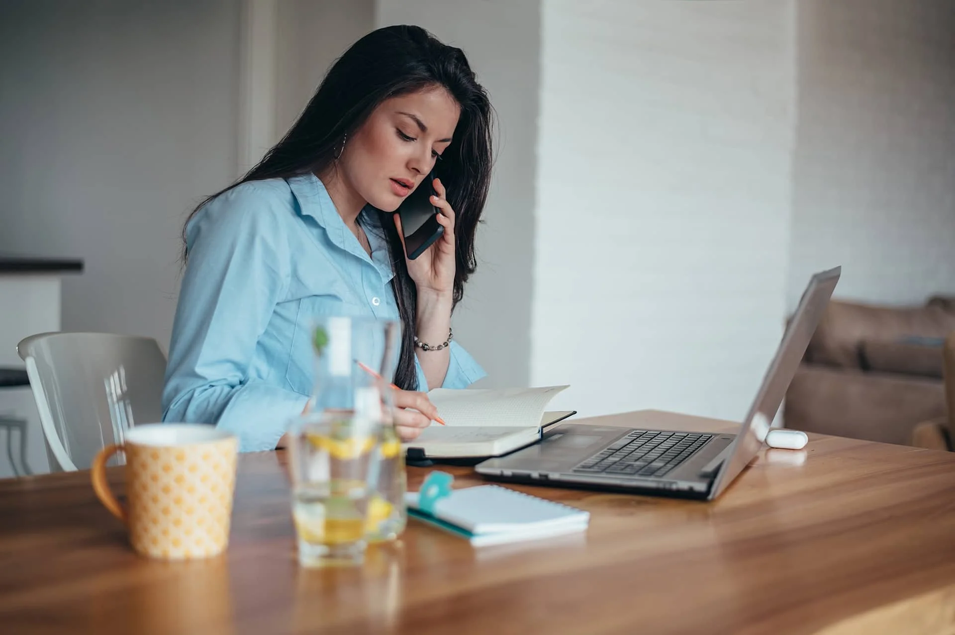 A woman sitting at a table using a laptop and phone, representing coordinating caregiving responsibilities from a distance.
