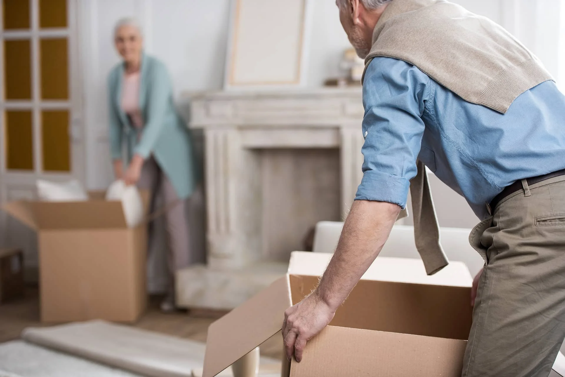 Man lifting a cardboard box while an older woman packs belongings in a living room during a home downsizing process.