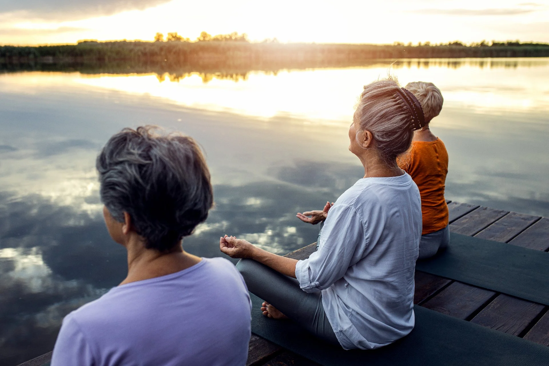 Three older women sitting on yoga mats on a wooden dock, meditating together by a calm lake at sunrise.