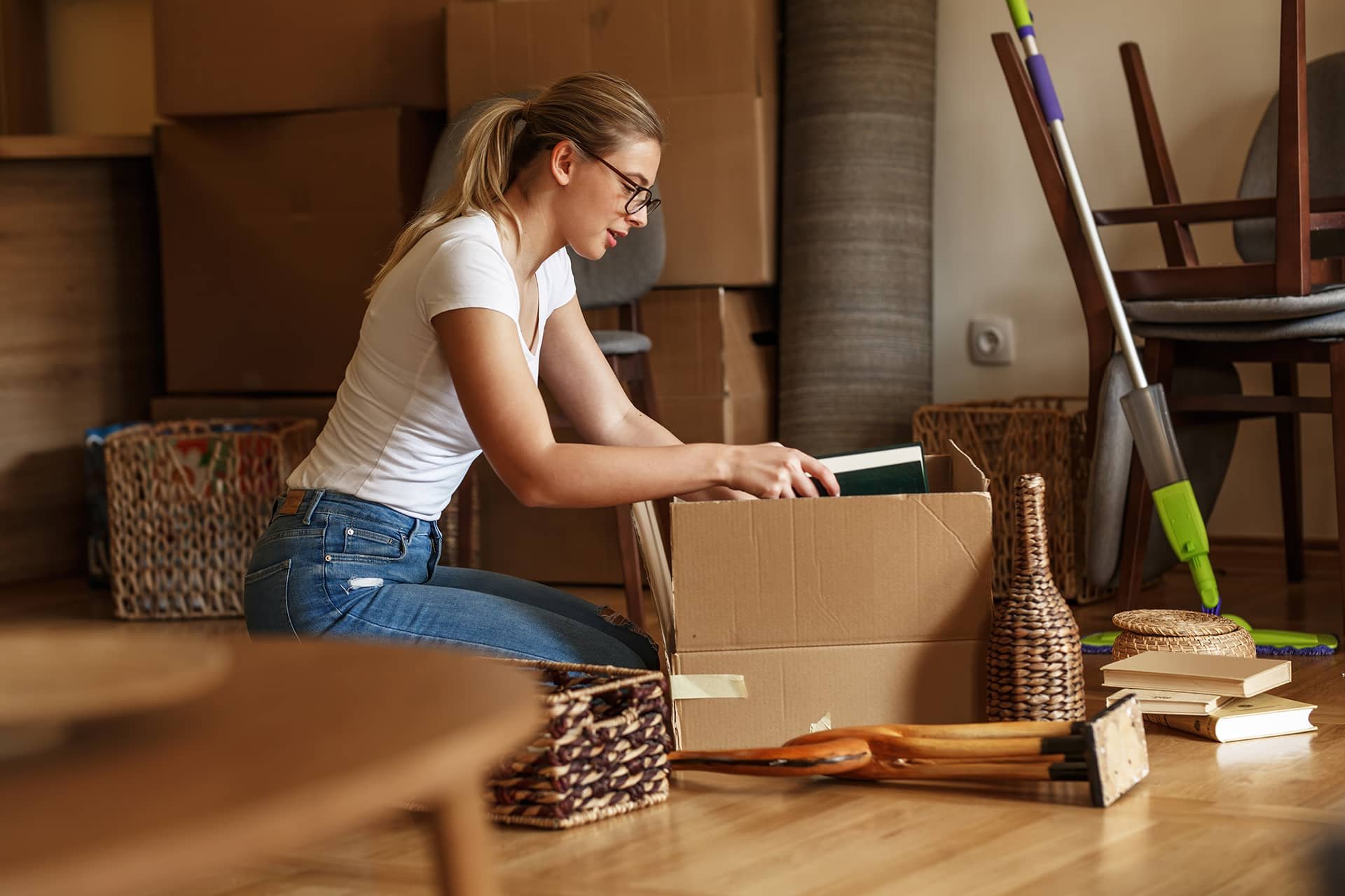 Woman sitting on the floor packing books and household items into a cardboard box during a home downsizing process.