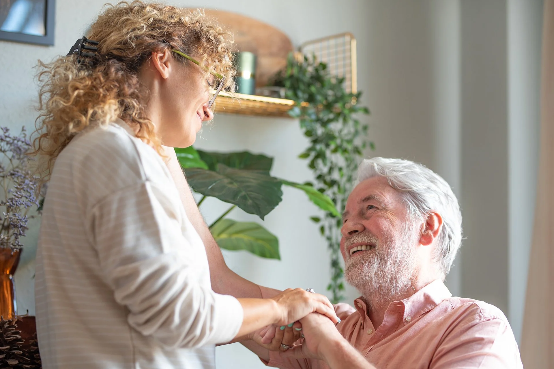 Caregiver smiling while holding hands with an older man during a warm conversation at home.