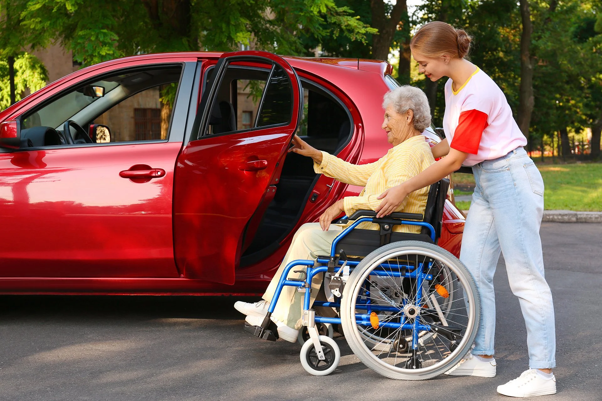 A young woman assists an older woman in a wheelchair beside an open car door, illustrating transportation support for someone with mobility needs.