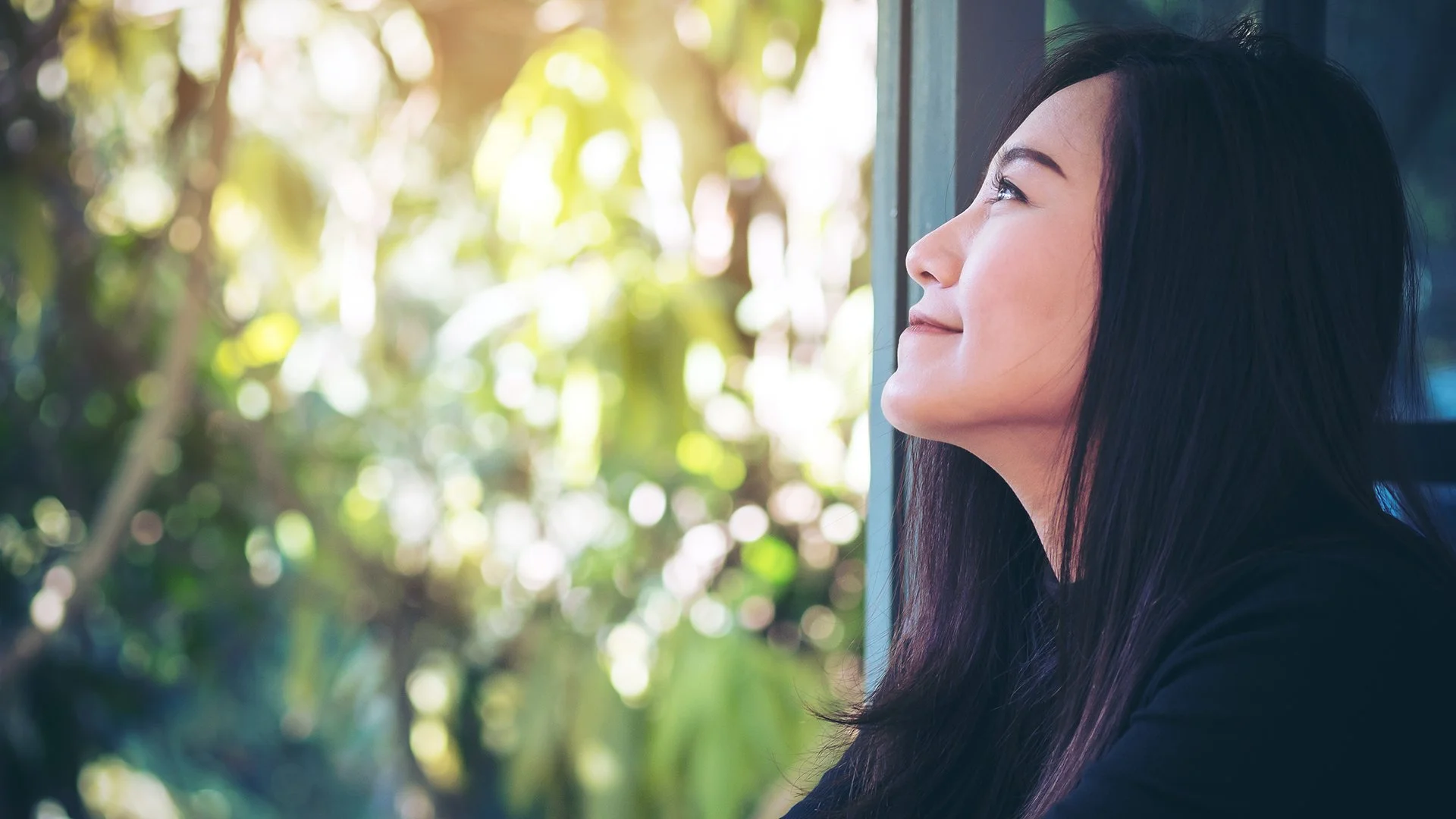 Woman sitting by a window, smiling gently as she looks upward with sunlight filtering through the trees outside.