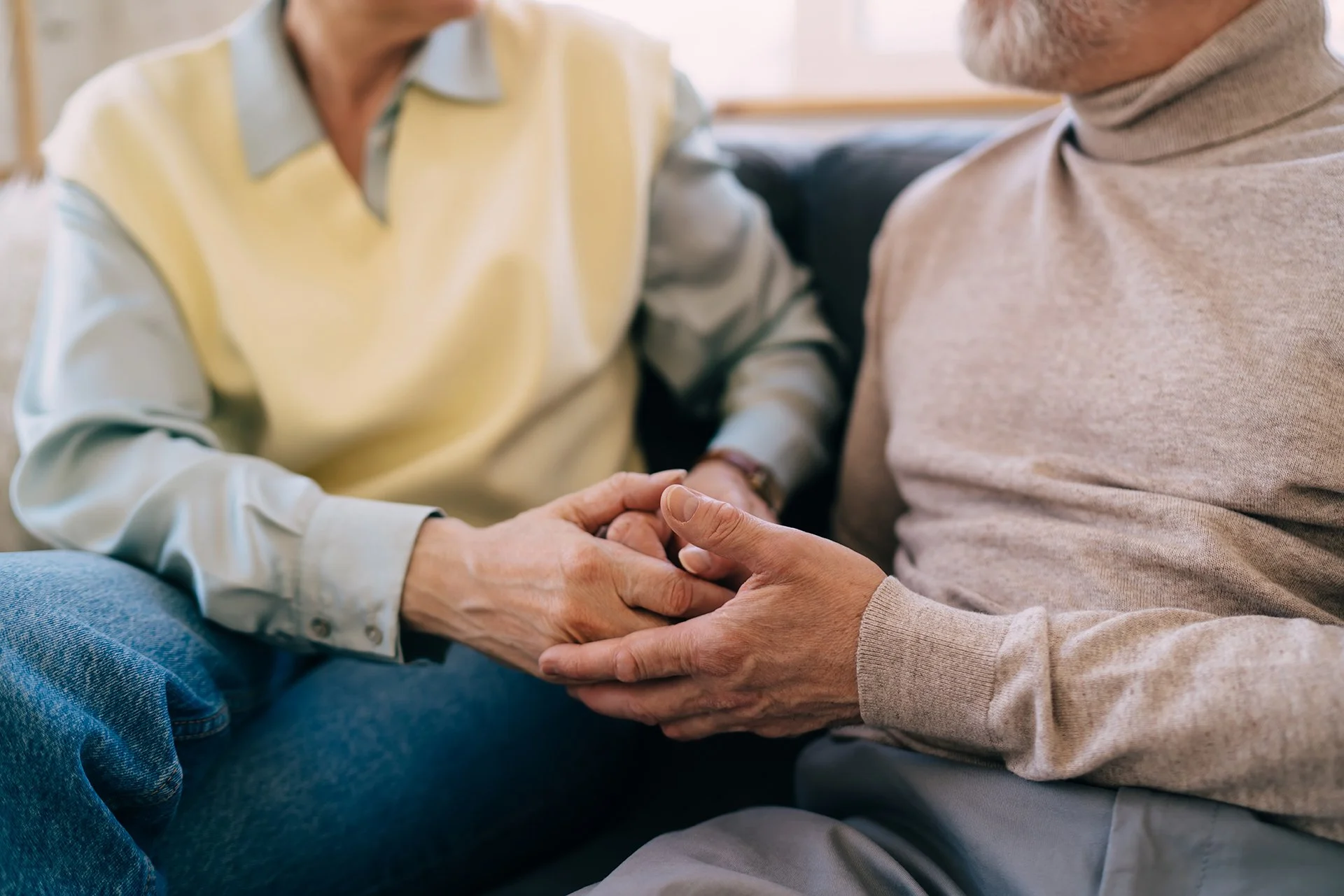 An adult child holds an aging parent's hands during a supportive conversation at home.