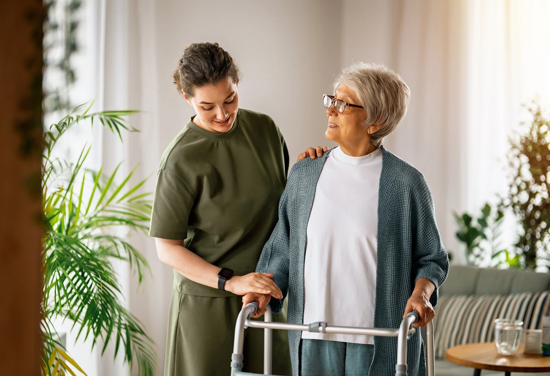 A caregiver assists an older woman using a walker while offering encouragement and support.