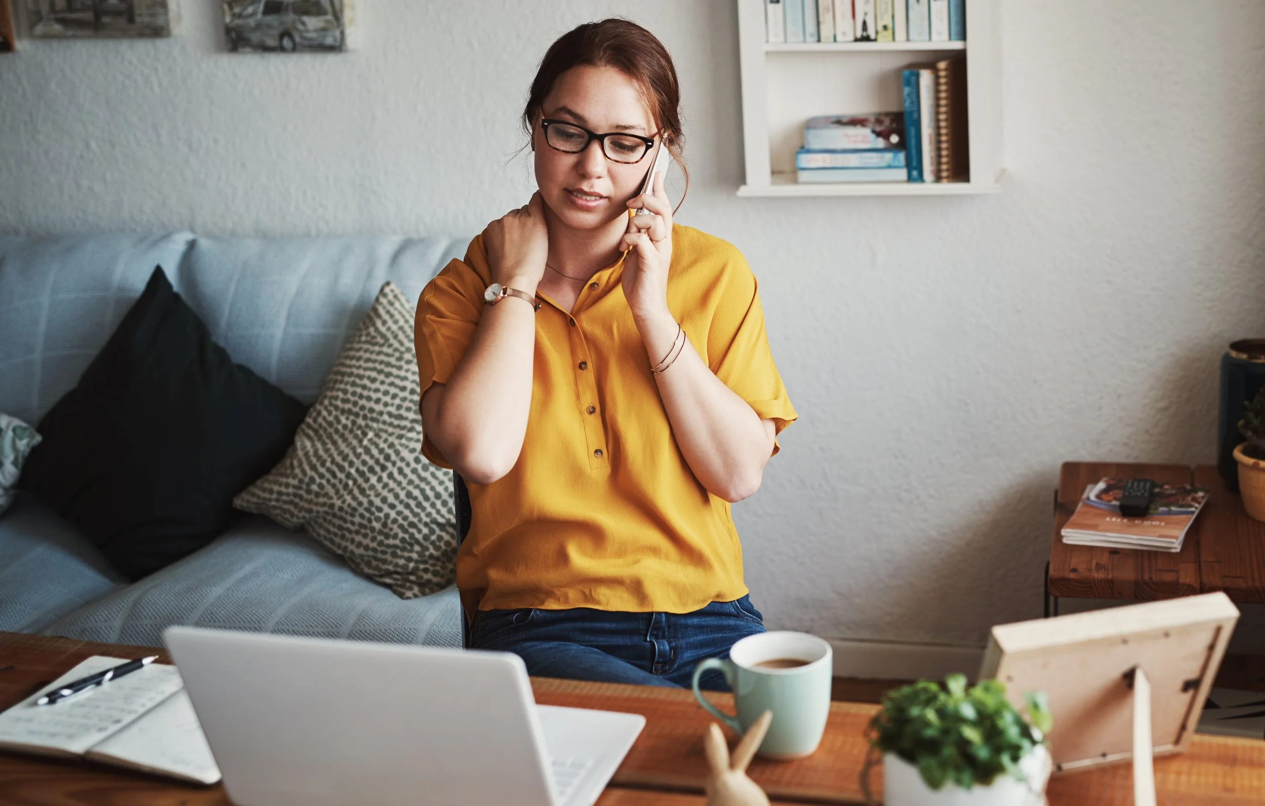 A woman talks on the phone while sitting at home with a laptop and notebook nearby, managing schedules and responsibilities.