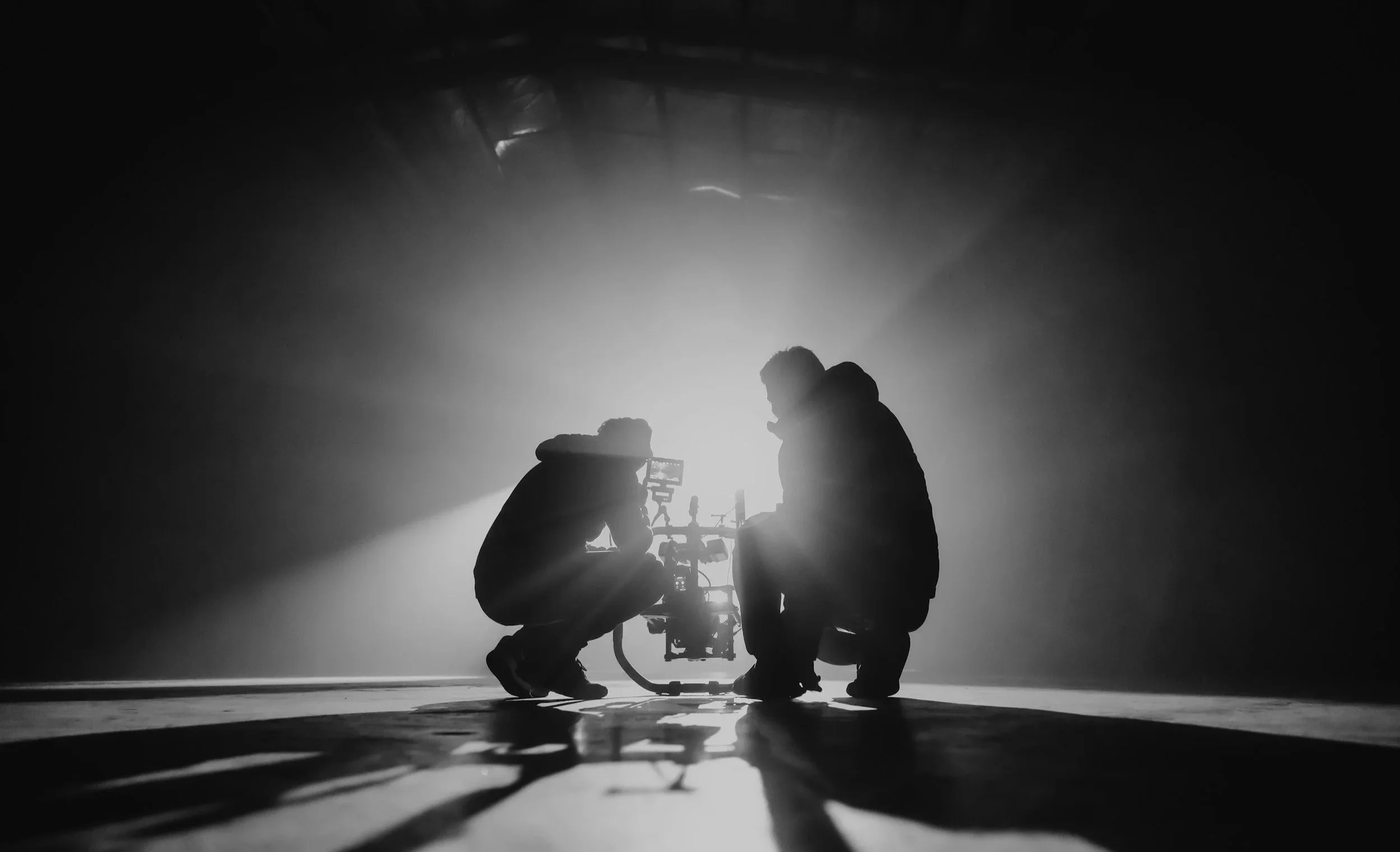 Two silhouetted people kneeling and working on film or photography equipment in a dark, foggy space with bright light shining from behind.