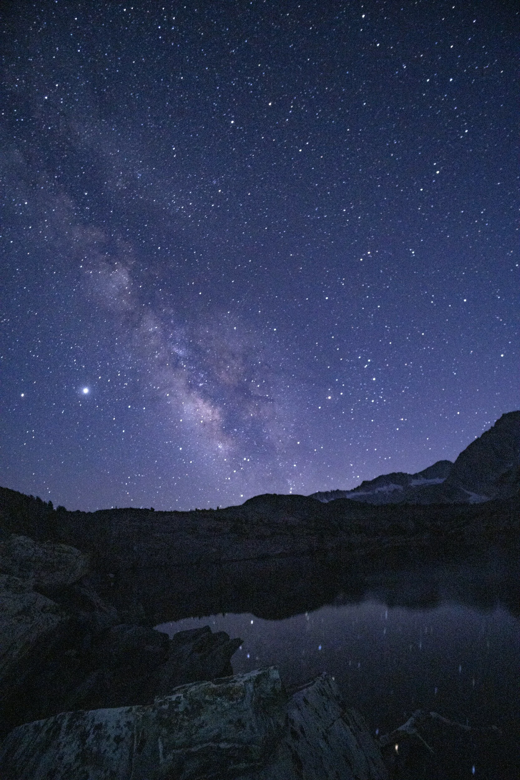 Night sky filled with countless stars, including the Milky Way galaxy, over a silhouette of rugged mountains and a calm reflective lake.