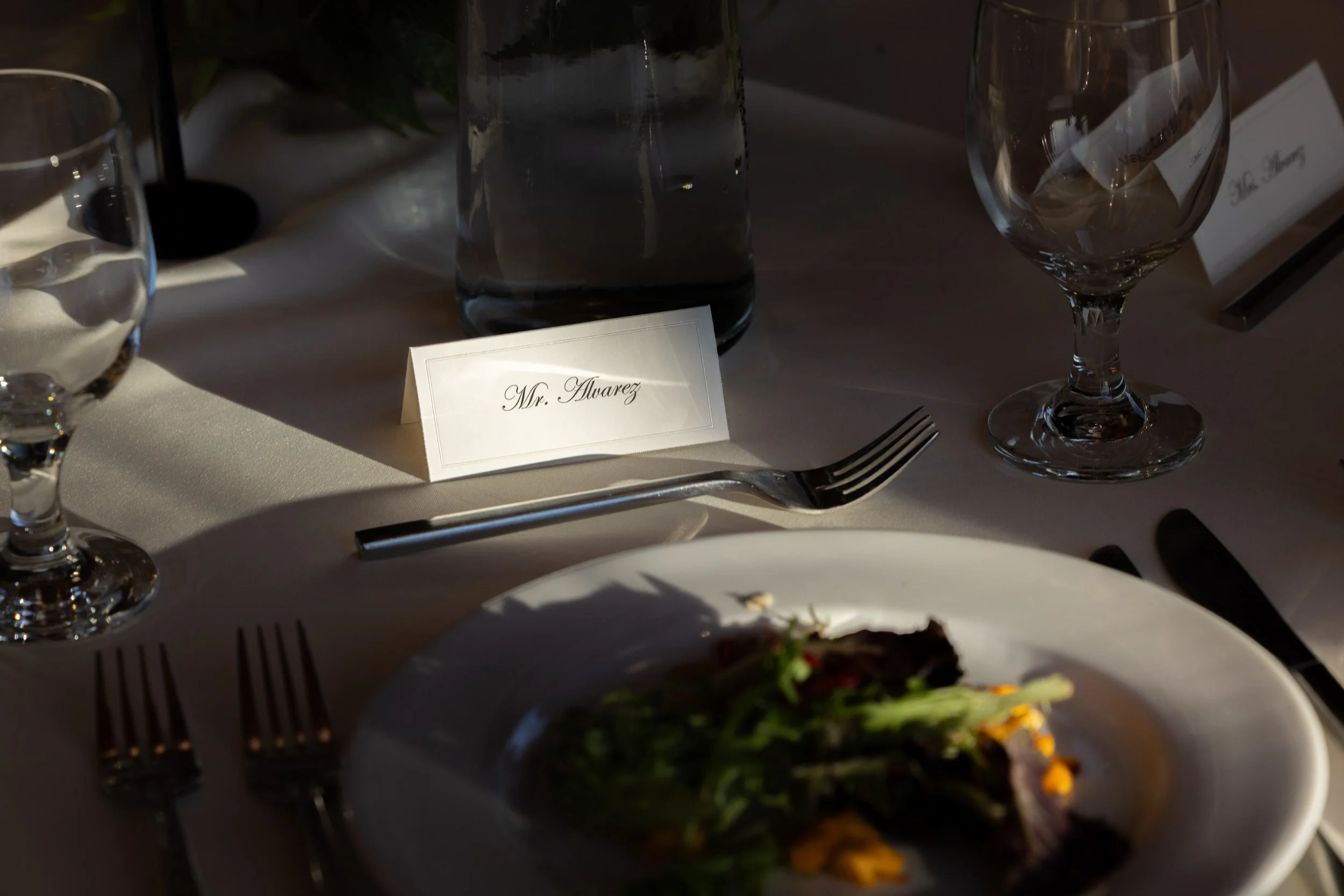 A table setting at a formal event featuring wine glasses, a water glass, a name card reading 'Mr. Alvarez,' a fork, and a plate with a salad. There is a tall water bottle and a second name card in the background.