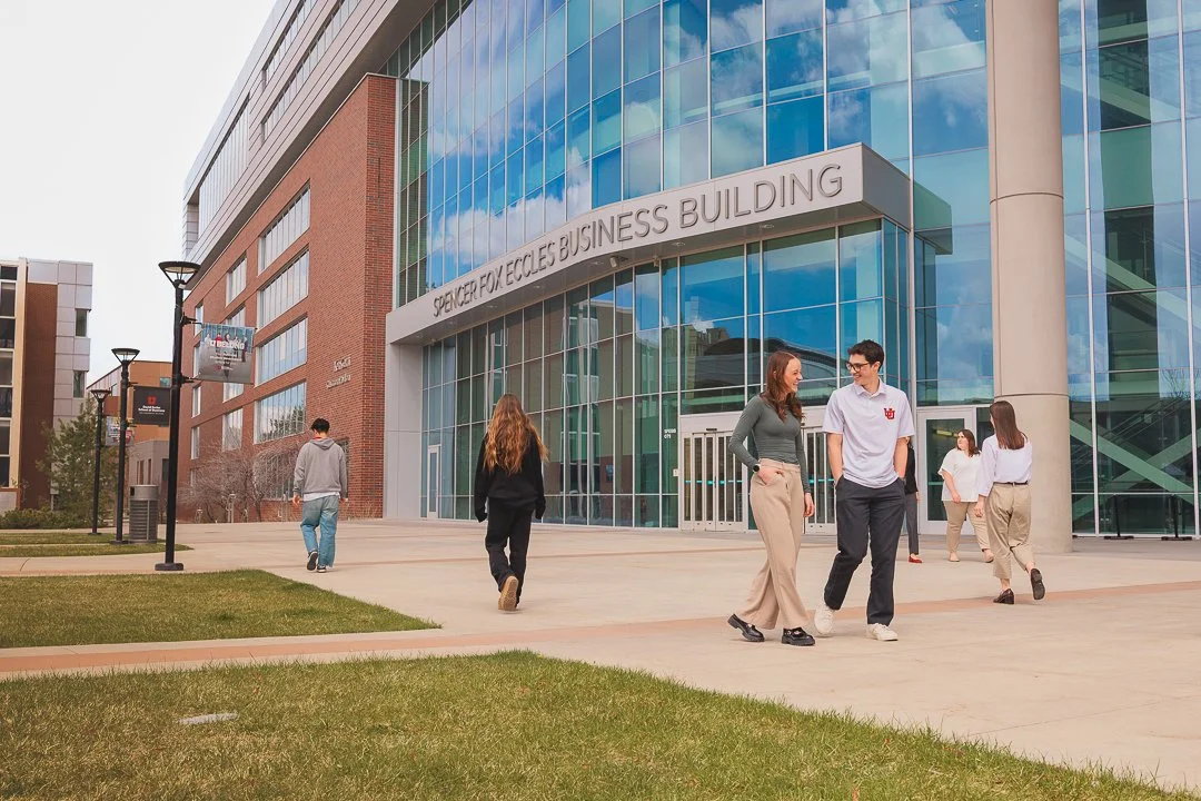 A modern glass and brick business building with a sign that reads 'Spencer Fox Eccles Business Building'. Several students are walking on the sidewalk outside the building.