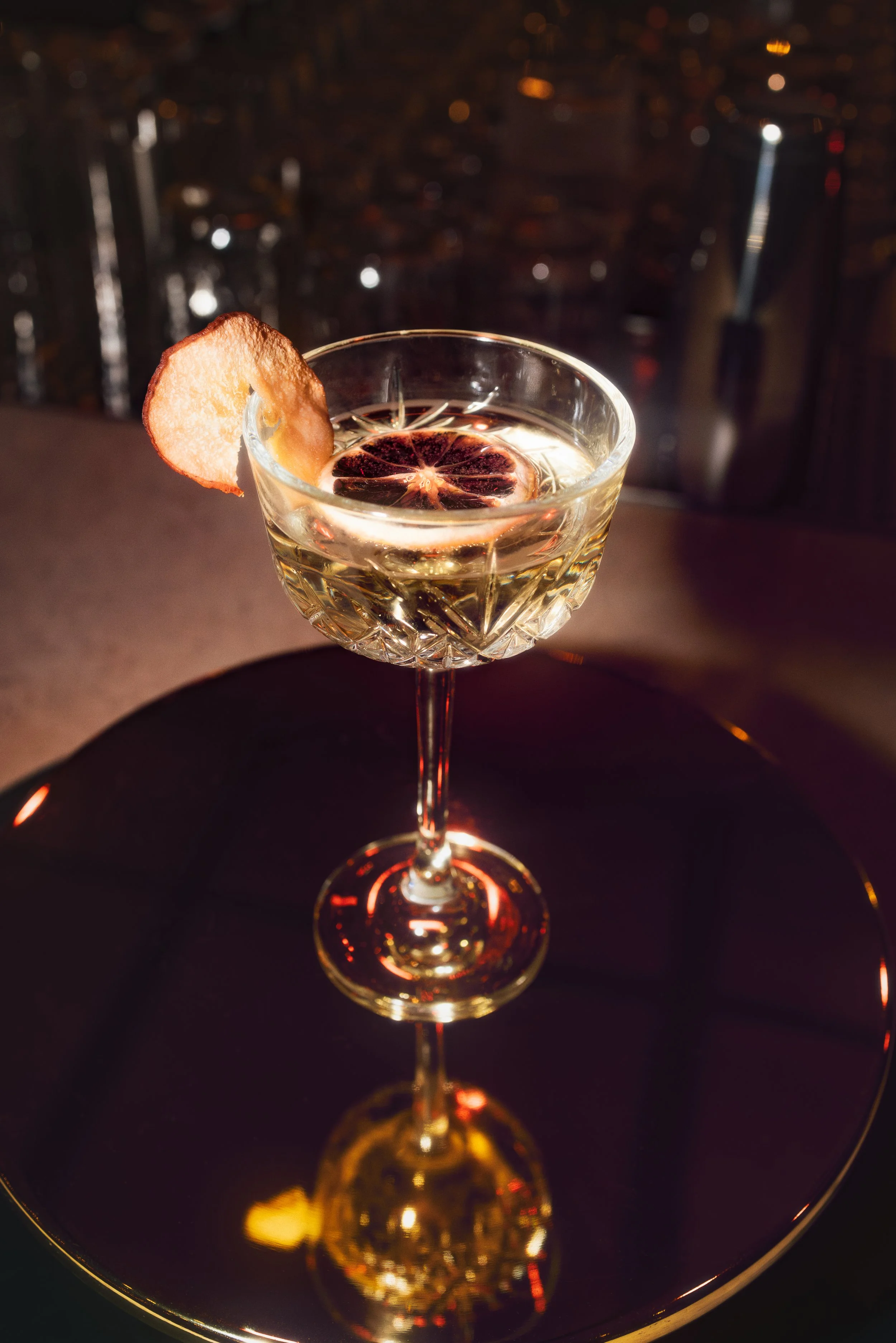 A crystal glass with a drink garnished with dried citrus slices and a plumb chip, set against a dark, reflective surface with city lights in the background.