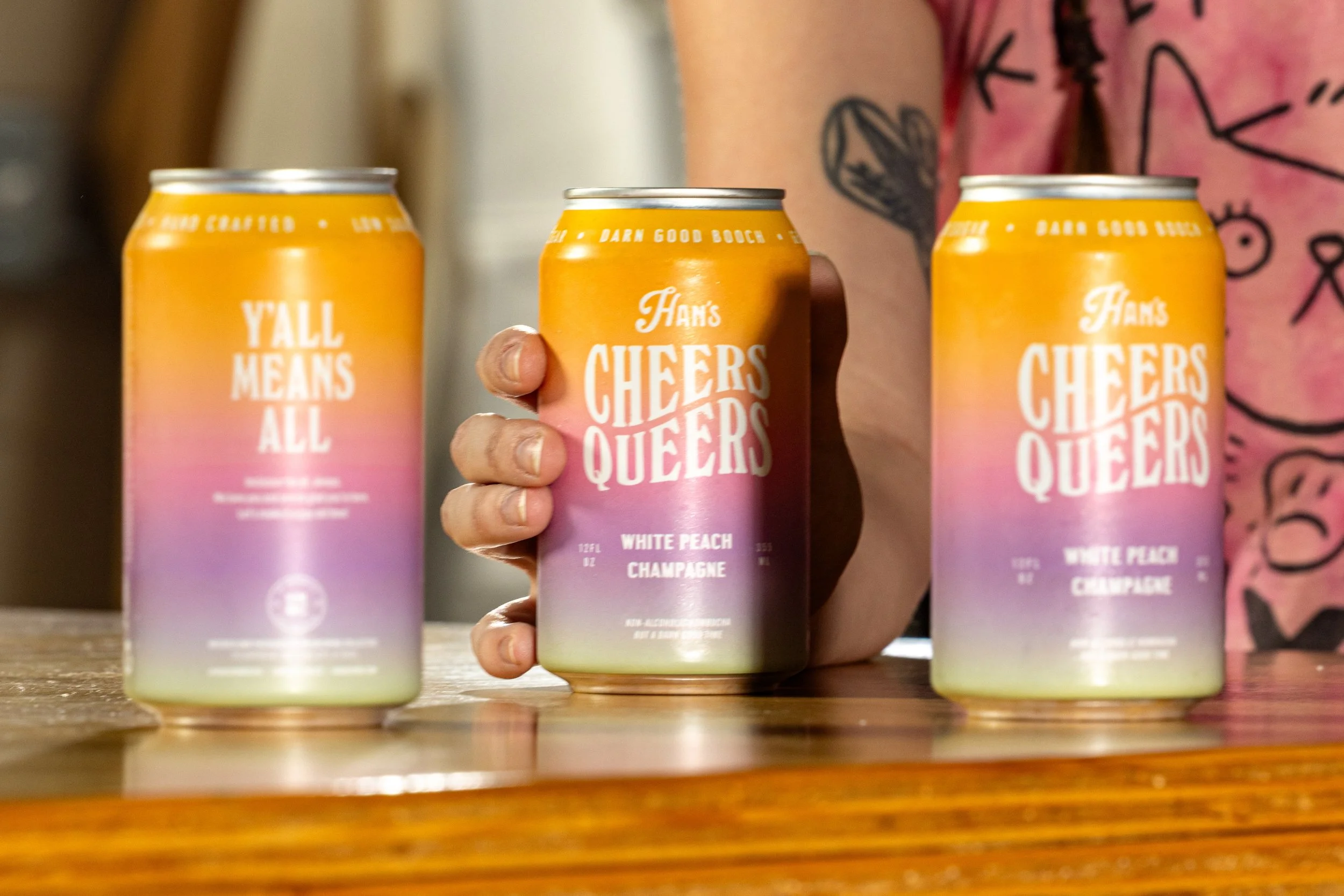 Three cans of Cheers Queers beverage with a gradient pink, purple, and yellow label with white text, held by a person with a tattooed arm, on a wooden table.