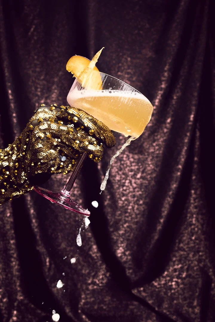 A hand wearing a black and gold sequin glove holding a champagne coupe glass with a light-colored cocktail and a plum garnish, against a dark textured background, with some liquid spilling from the glass.