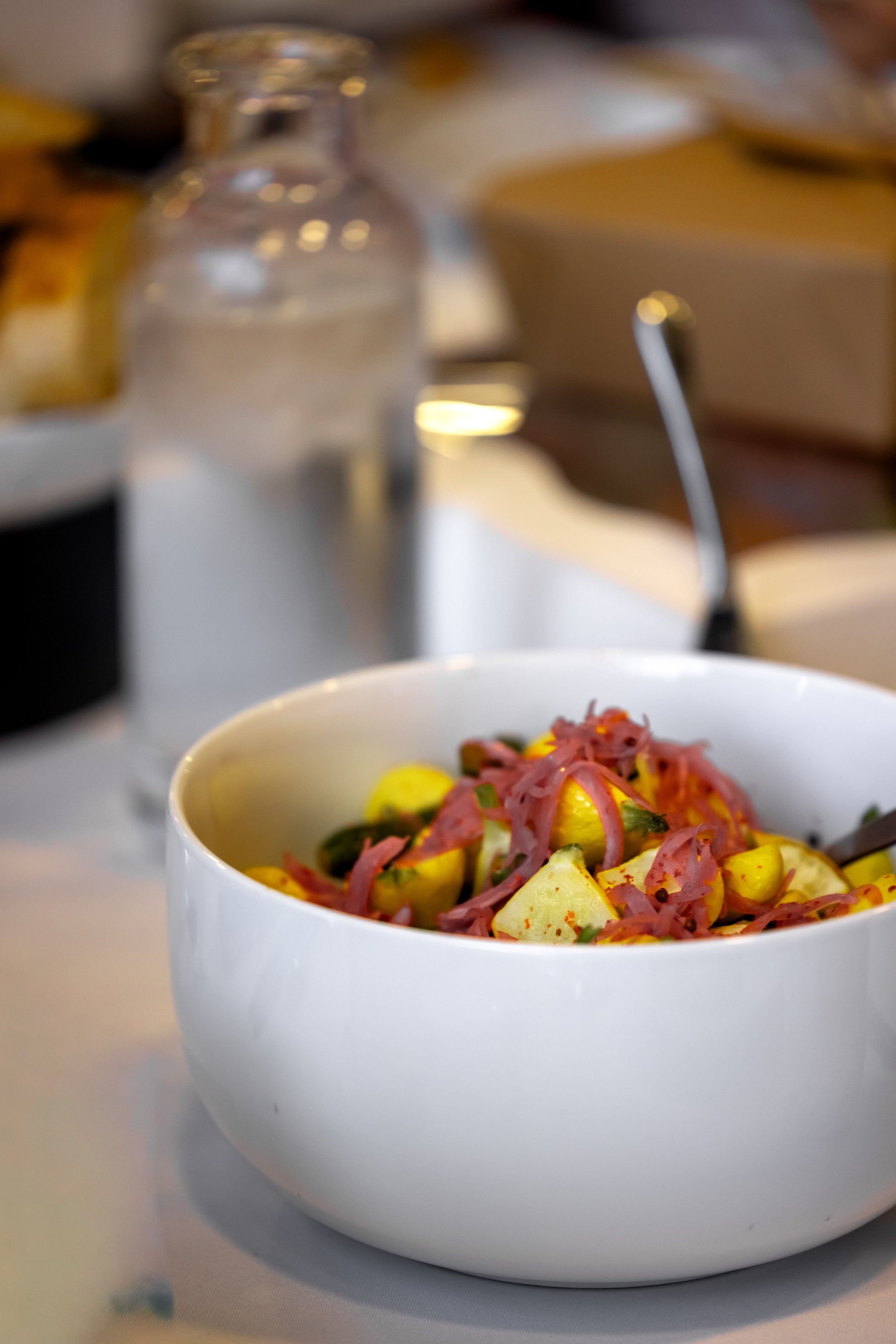A white bowl filled with a colorful salad containing yellow potatoes, green herbs, and shredded pink meat, set on a white surface with a blurred background.