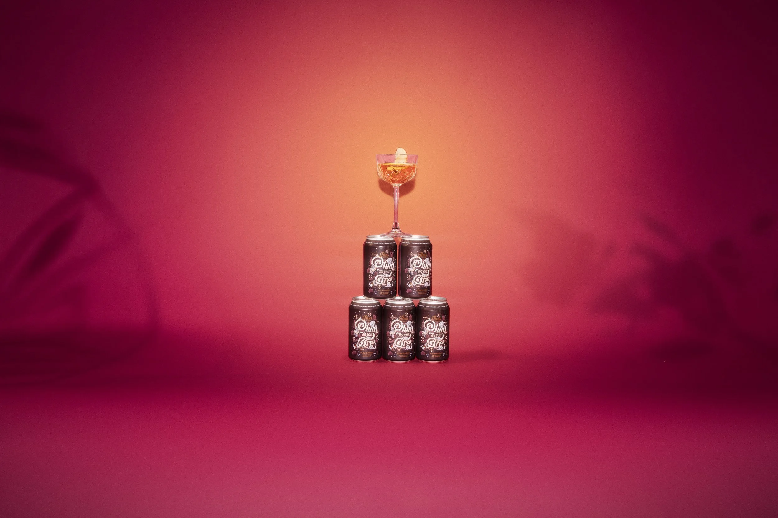 A pyramid of eight cans with a glass on top filled with a pink cocktail topped with a white decorative straw. The cans have a festive design and text that says "Plumb as you Are". The background is a gradient pink and orange.