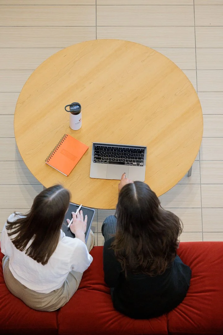 Two women sitting on a red couch at a round wooden table. One woman is using a tablet, the other is using a laptop. Also on the table are a water bottle, an orange notebook, and a tabletop ventilator.