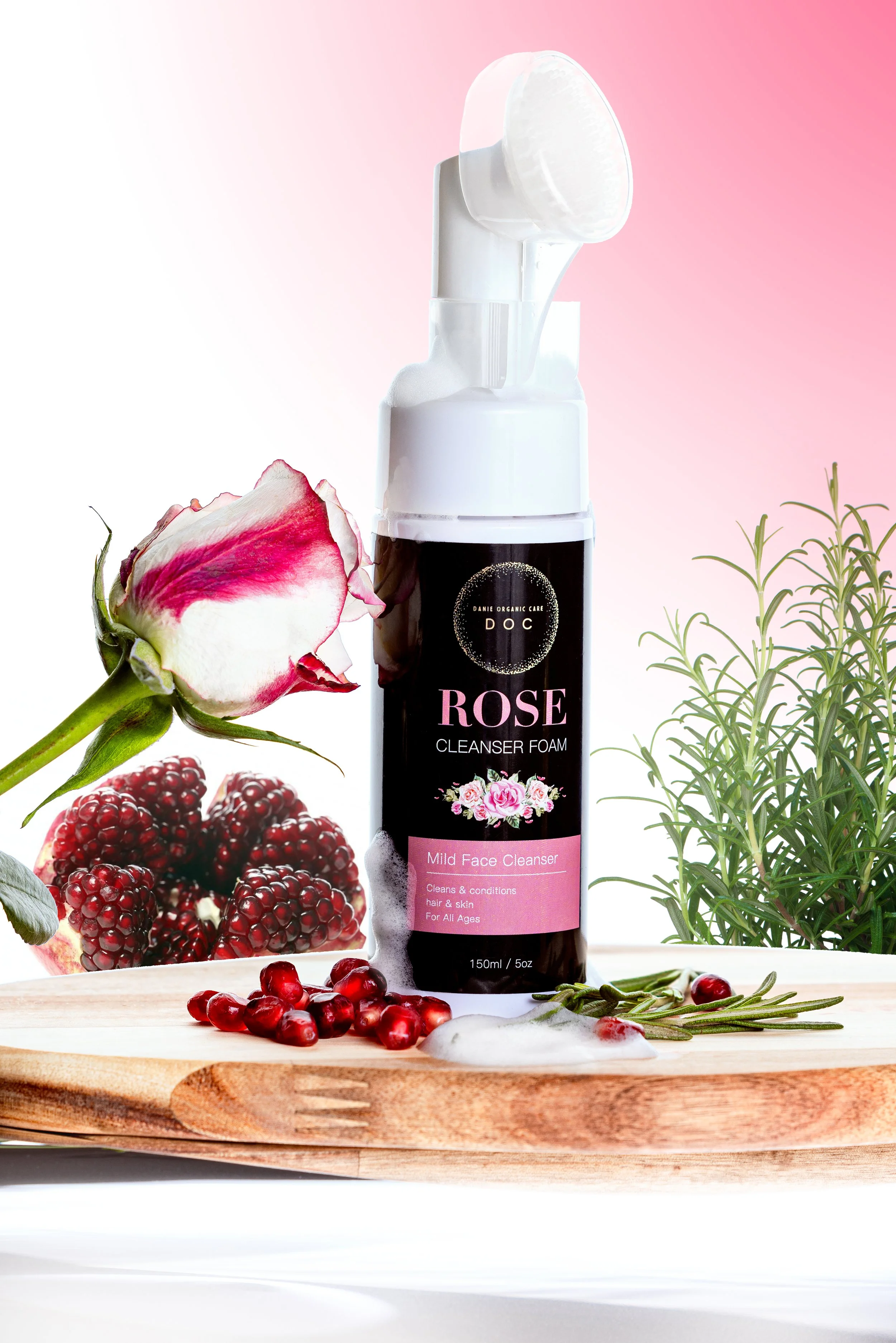 A bottle of rose cleanser foam on a wooden surface with pomegranate seeds, a rose flower, and green herbs around it, set against a pink gradient background.