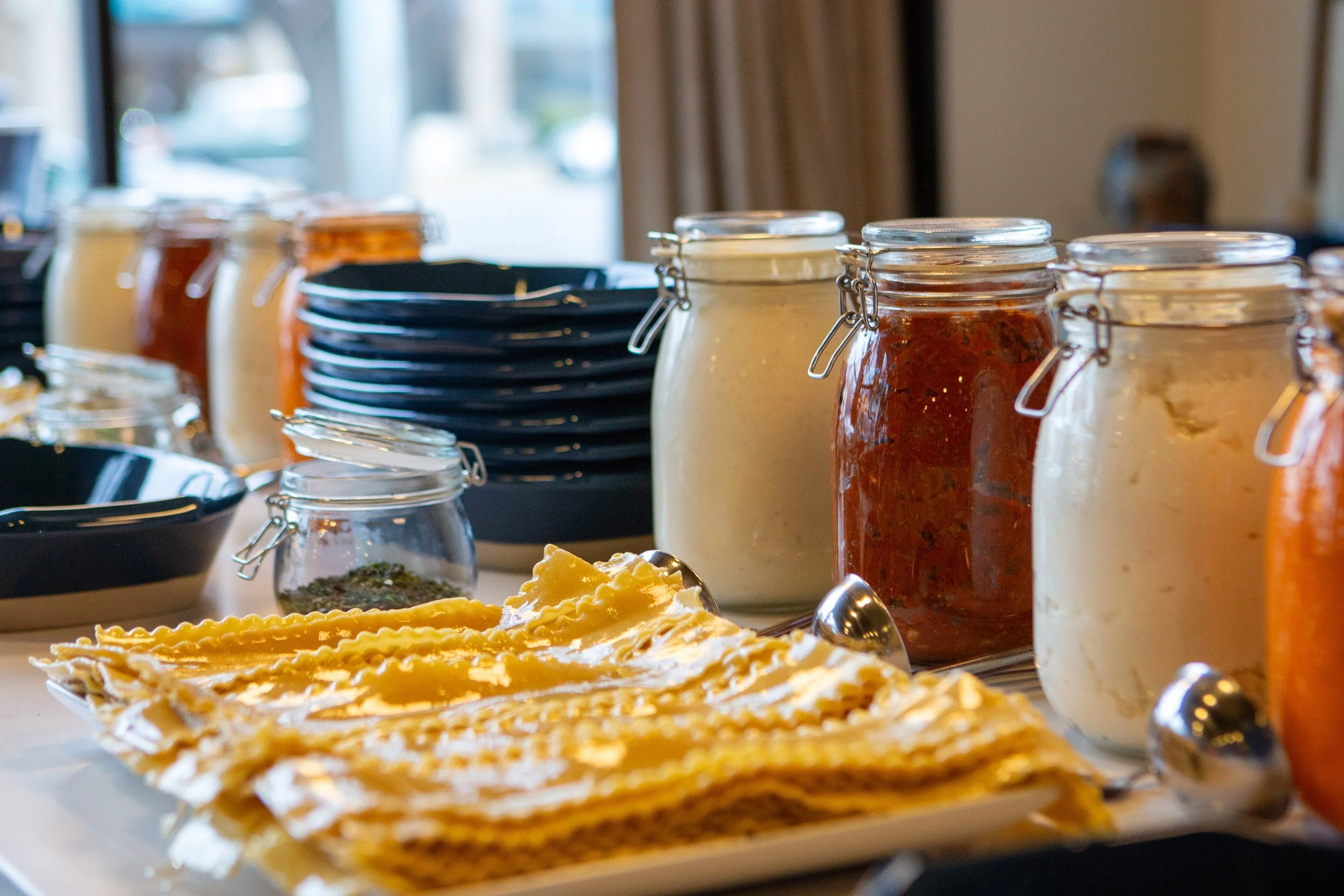 A close-up of a build-your-own lasagna in the foreground and various jars of different sauces or spreads behind, along with stacked black plates and small bowls, with a window and curtains in the background.
