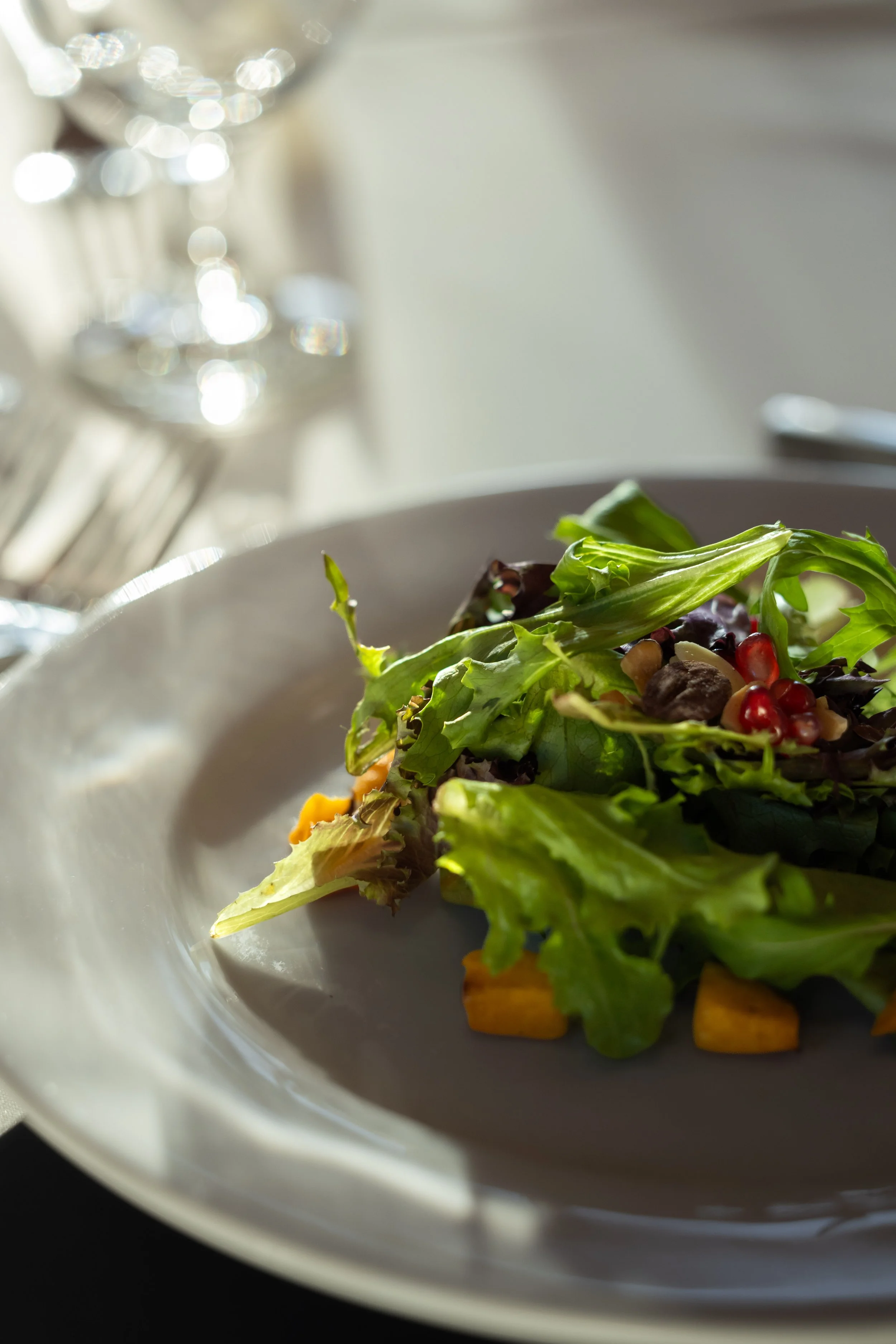 Close-up of a fresh salad with mixed greens in a white bowl on a table. In the background, there is a glass and silverware.