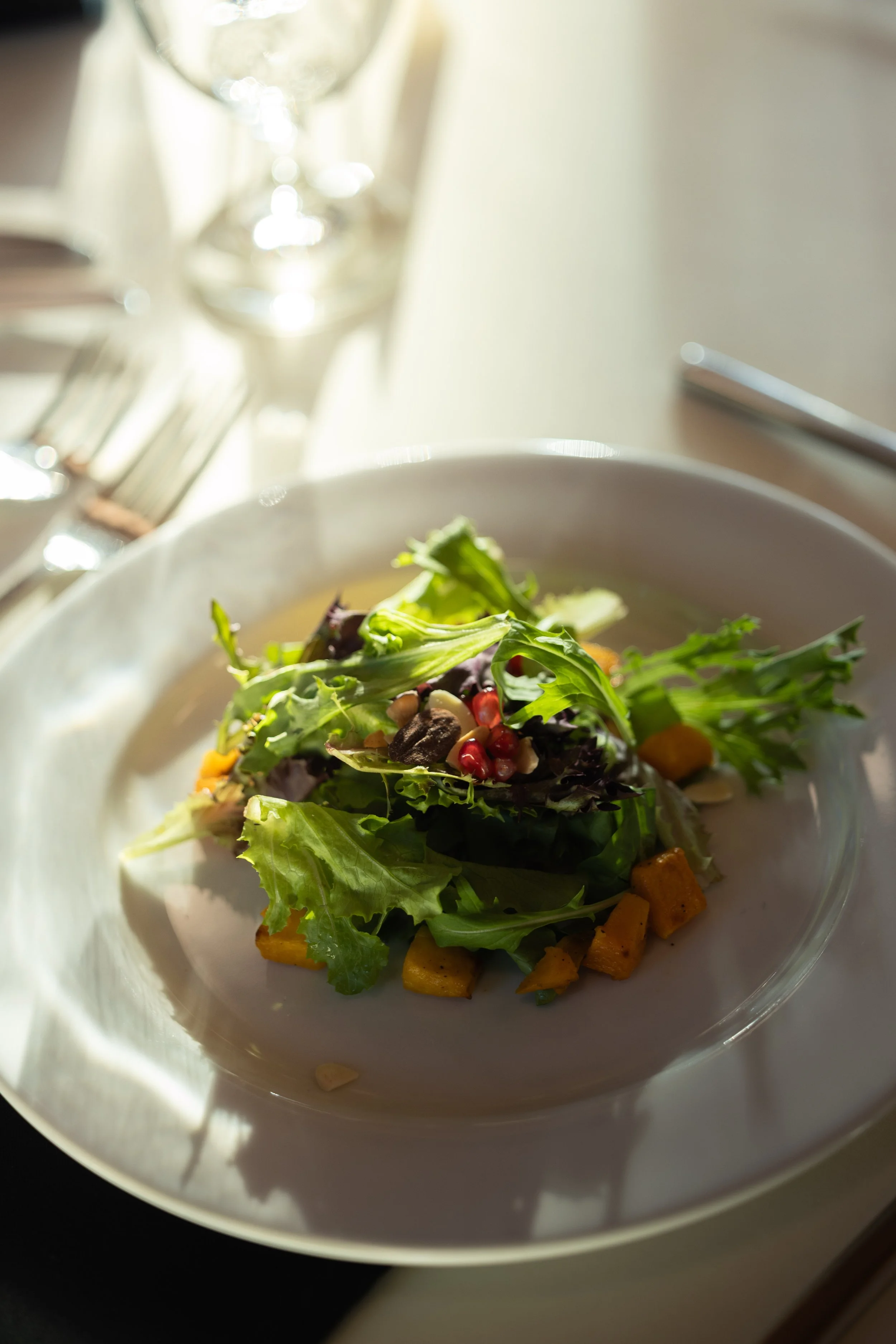 A close-up of a salad with mixed greens, diced butternut squash, pomegranate seeds, and nuts in a clear glass bowl on a white table, with a blurred glass of white wine and silverware in the background.