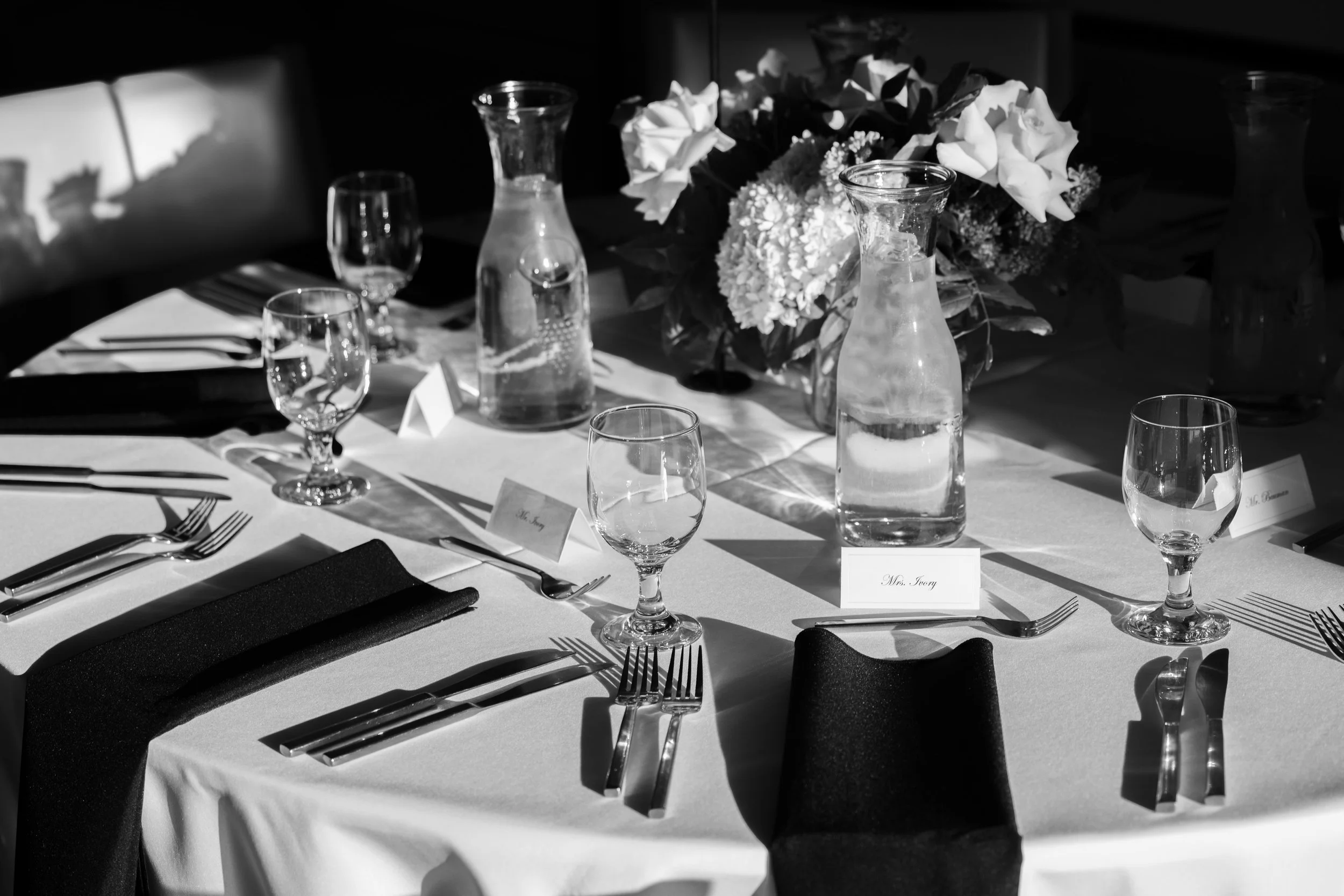 A black and white photograph of a formal dining table set with glassware, cutlery, napkins, and a flower centerpiece with white roses and other flowers.