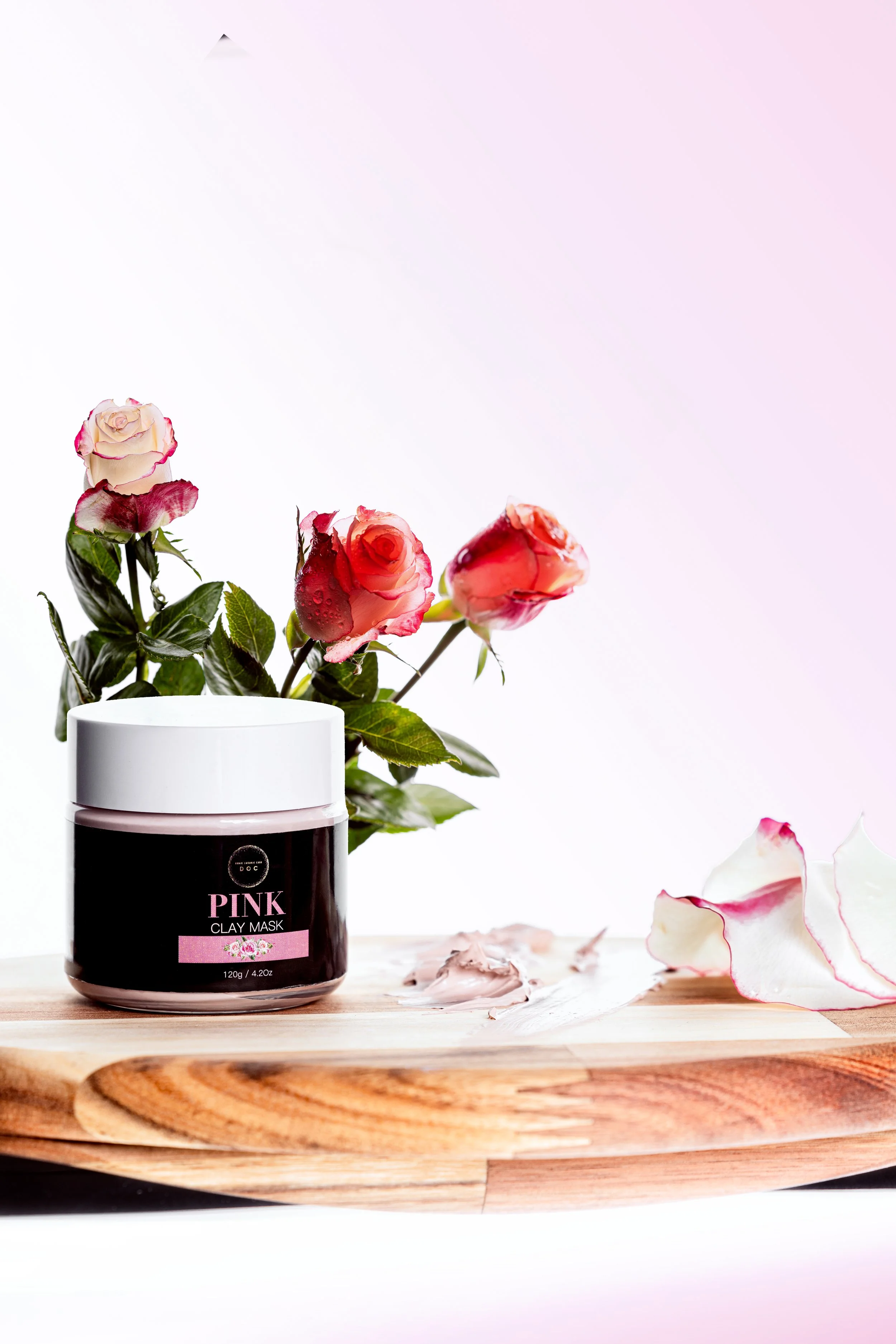 A black jar labeled 'Pink Clay Mask' with pink flowers in a white vase placed on a round wooden tray with scattered flower petals and clay mask residue.