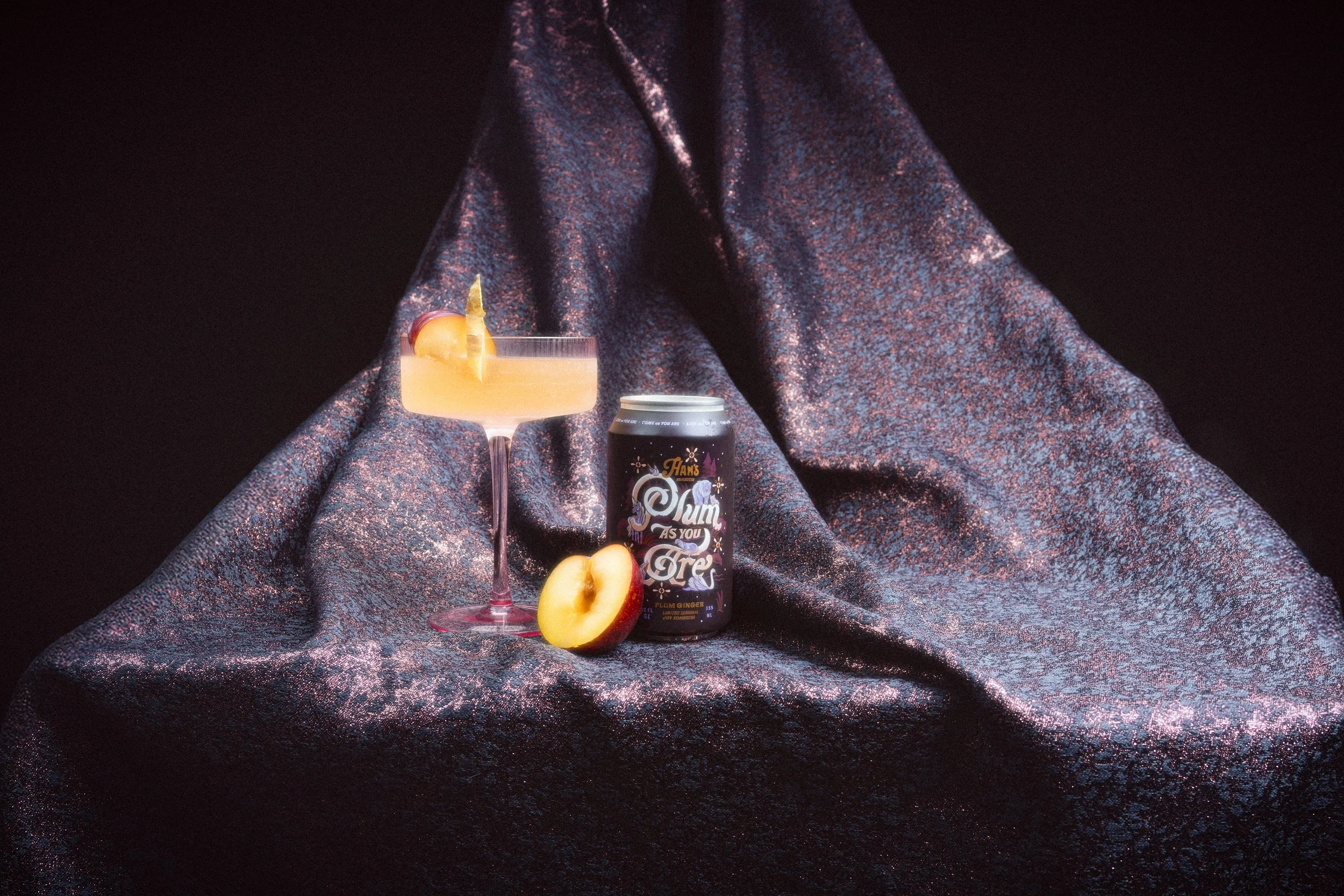 A cocktail glass with a peach slice garnish, a can of Plum Ginger drink, a halved plumb, and a dark, glittery fabric background.
