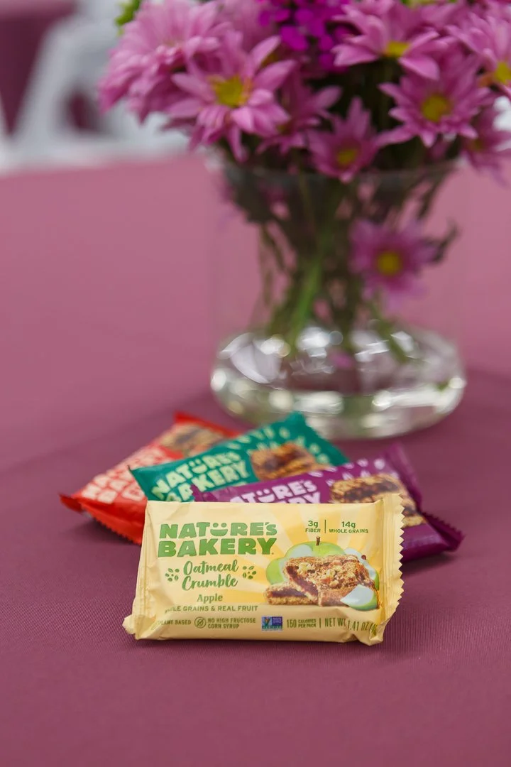 Three Nature's Bakery oat bar packages in front of a clear glass vase filled with pink flowers on a pink surface.