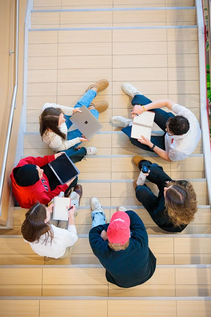 Group of six young people sitting on wooden stairs in a circle, engaged in a discussion or study session with notebooks, tablets, and water bottles.